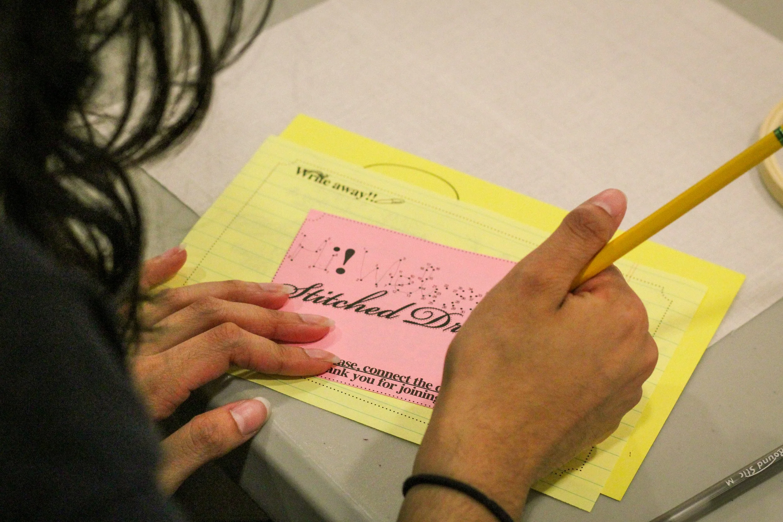 A person with dark curly hair is writing on a pink and yellow paper titled 'Stitched'. The paper has text, design, and a handwritten part, held down with one hand while using a yellow pencil with the other.
