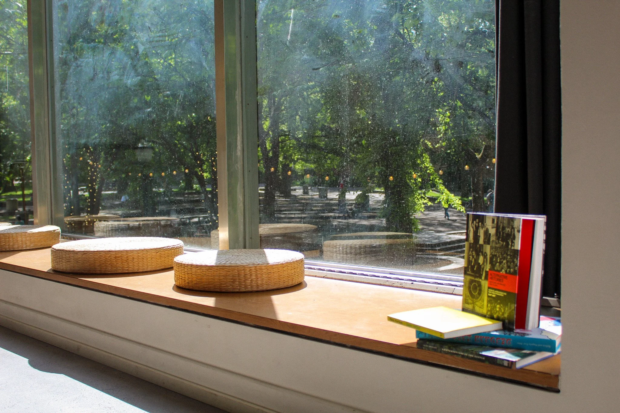 Sunlit window seat with three round woven cushions and a stack of books on the windowsill, overlooking a park with trees and string lights outside