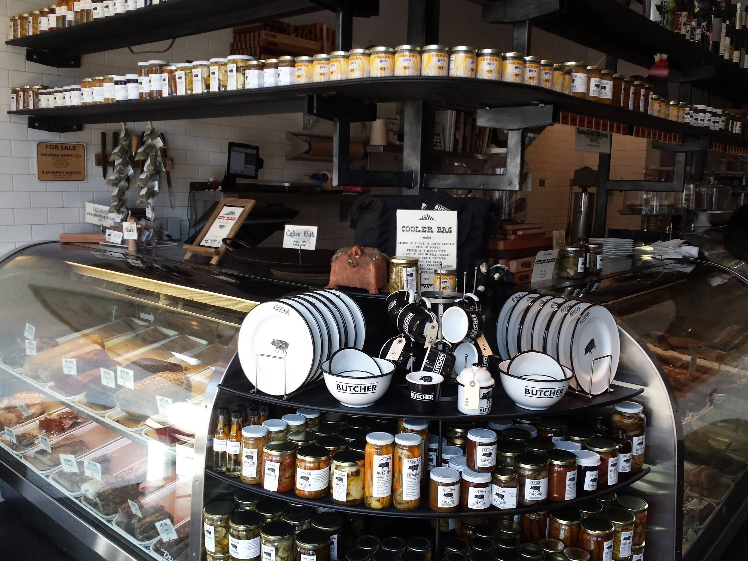 Interior of Cochon Butcher with a glass display case, jars of condiments, white tiled walls, and shelves of packaged goods.