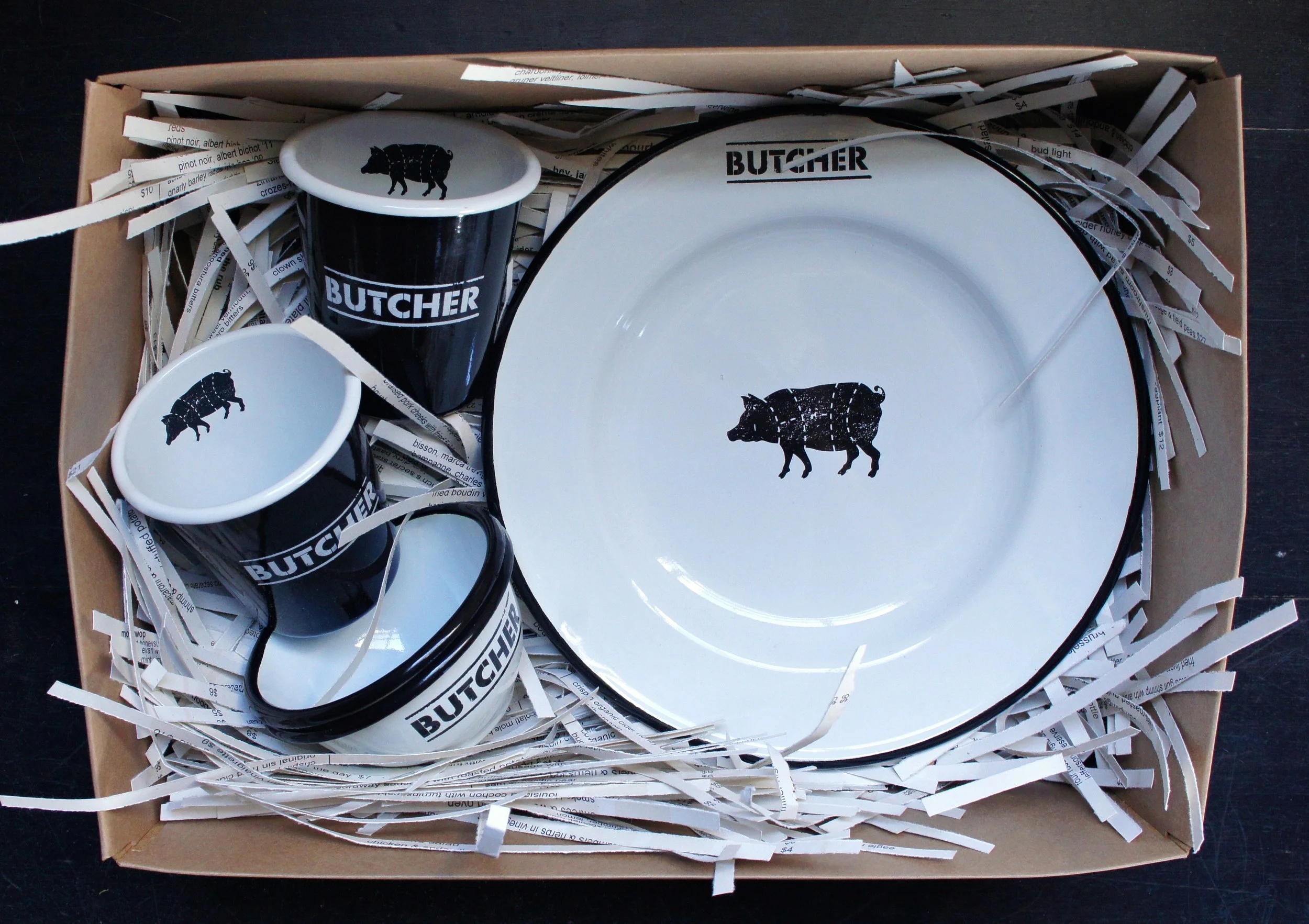 Boxed Cochon Butcher enamelware gift set with white plates printed with black pig silhouettes and black branded mugs.