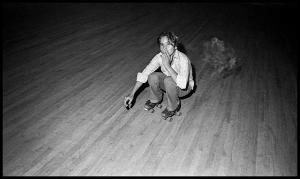 Black-and-white Bill Yates photograph from Sweetheart Roller Skating Rink showing a young skater crouched on the rink floor smoking, used as visual inspiration for the raw, offbeat tone of More Salt, More Acid.