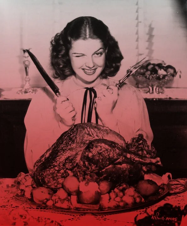 Red-tinted archival photograph of Rita Hayworth seated behind a roast turkey and fruit on a Thanksgiving table.