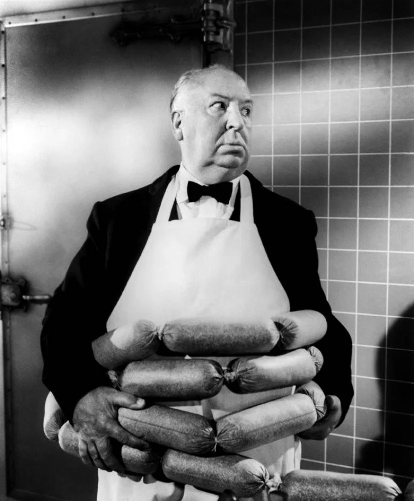 Black-and-white photograph of Alfred Hitchcock in a tuxedo and apron holding an armful of sausages, used as visual inspiration for the deadpan food humor and surreal tone of More Salt, More Acid.