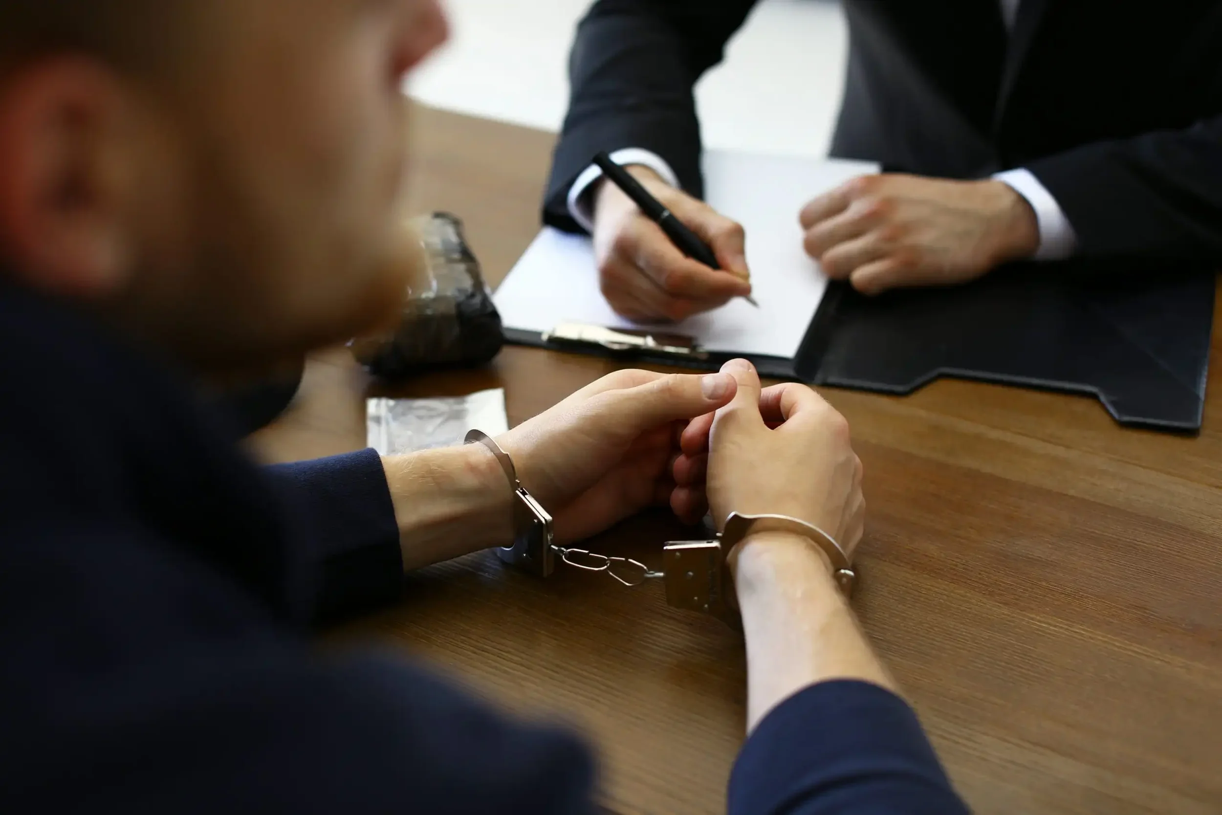 A person in handcuffs sitting at a wooden table with another person in a suit writing on a notepad.