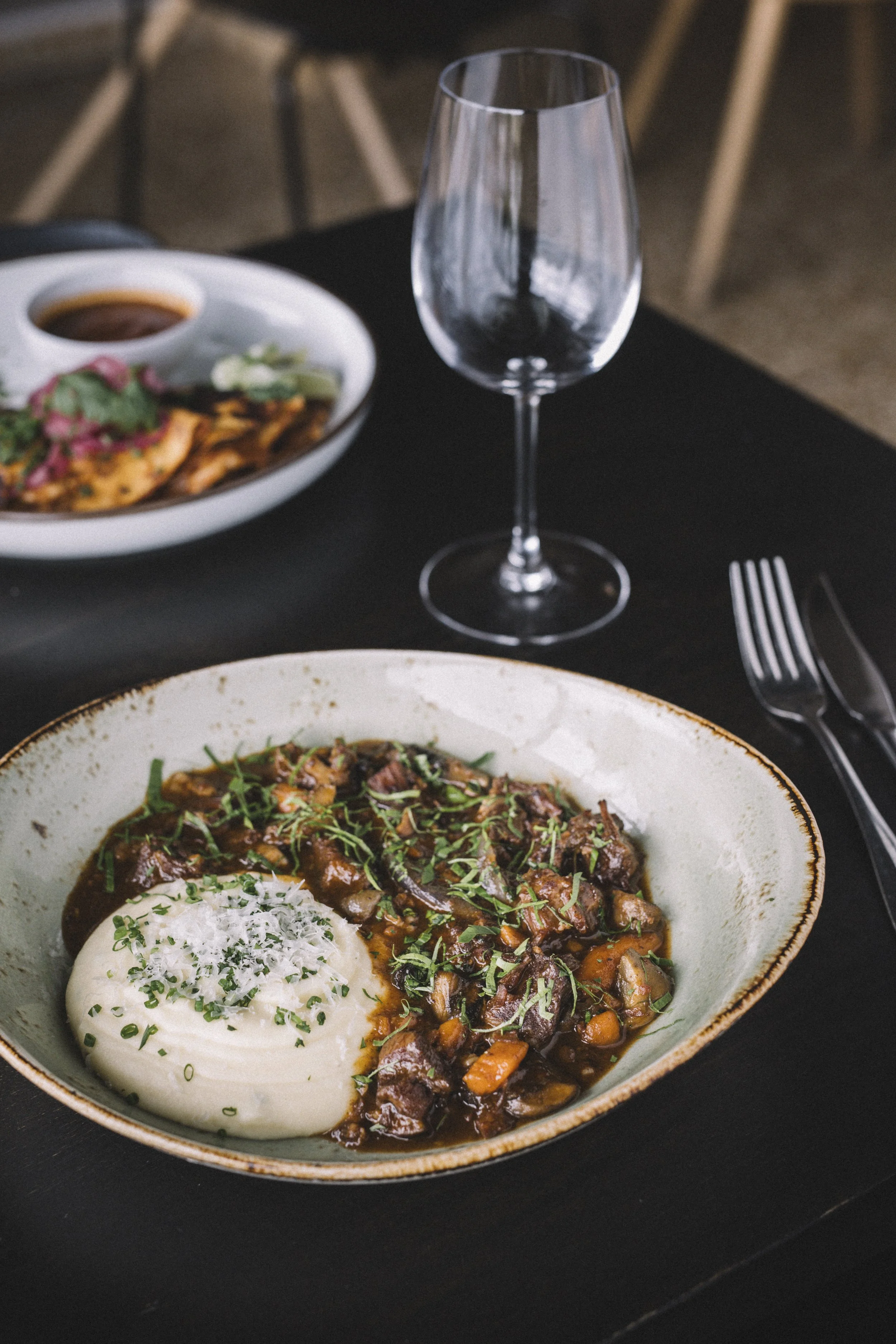 En tallerken med kjøttgryte og potetmos, pyntet med urter, på et svart bord ved siden av et tomt vinglass og bestikk. I bakgrunnen en skål med mat og saus.