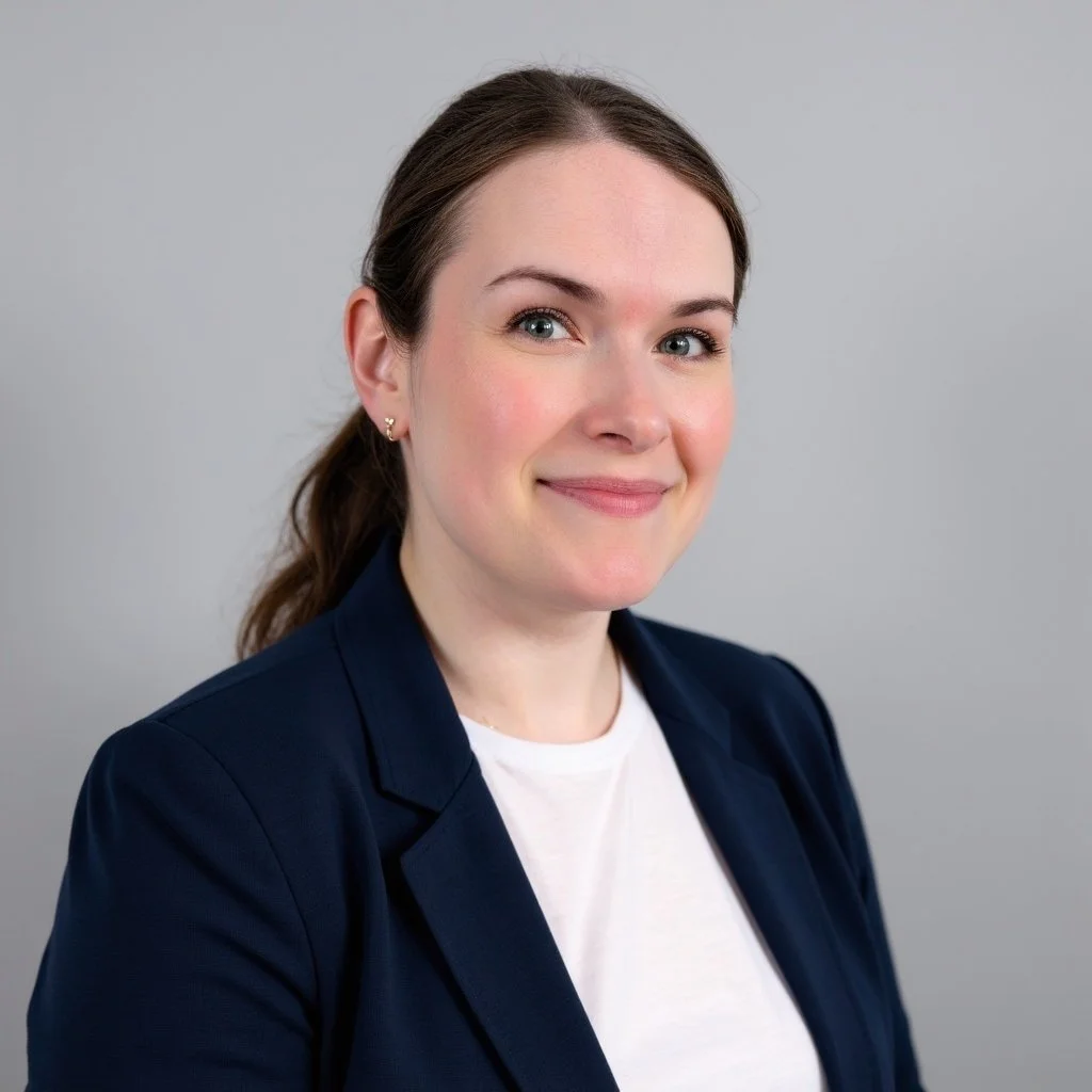 A portrait of a woman with brown hair tied back, wearing a navy blazer and a white top, smiling slightly, against a plain light gray background.