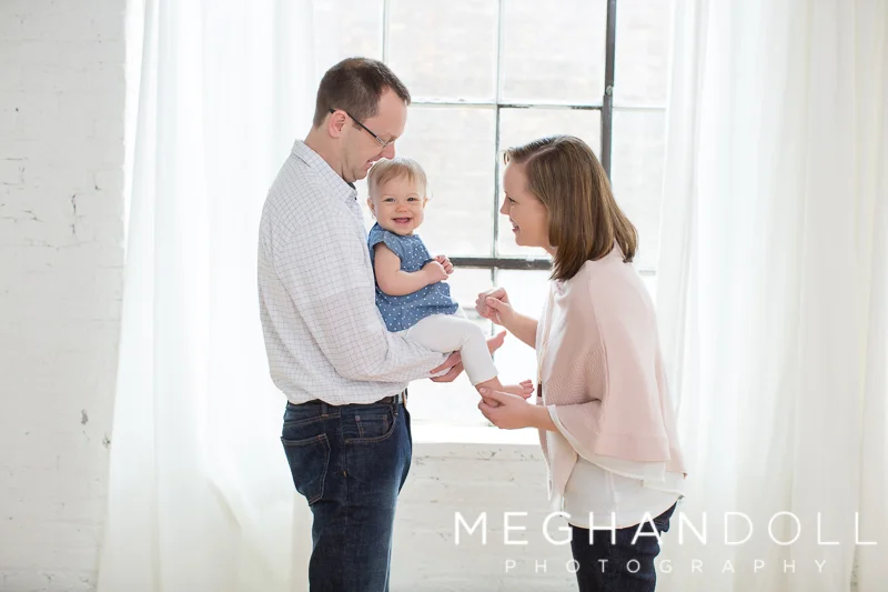 mom-and-dad-play-with-one-year-old-girl-by-window