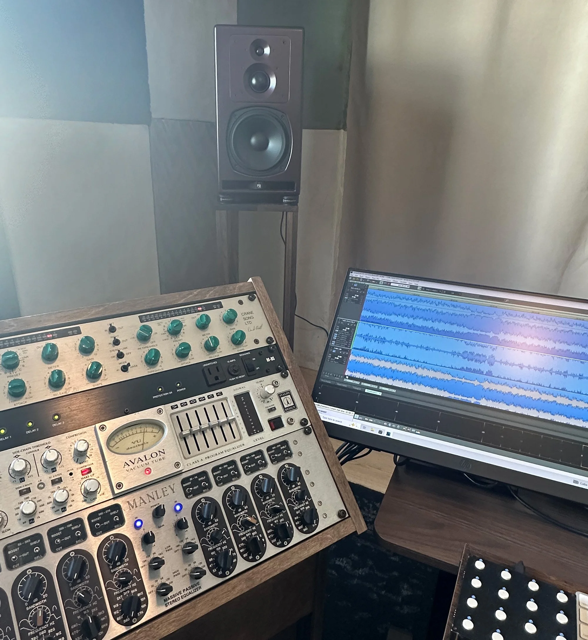 Music studio with a computer monitor displaying an audio editing software, a speaker on a stand, and a wooden audio mixing console with various knobs and controls.