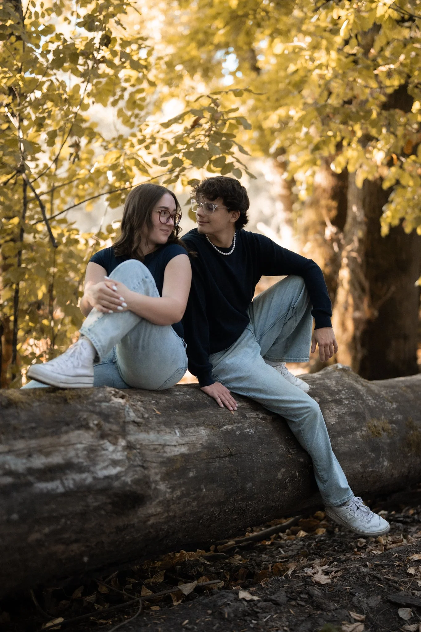 Two young women with glasses sitting on a fallen tree trunk amidst golden autumn leaves, looking at each other affectionately in a forest.