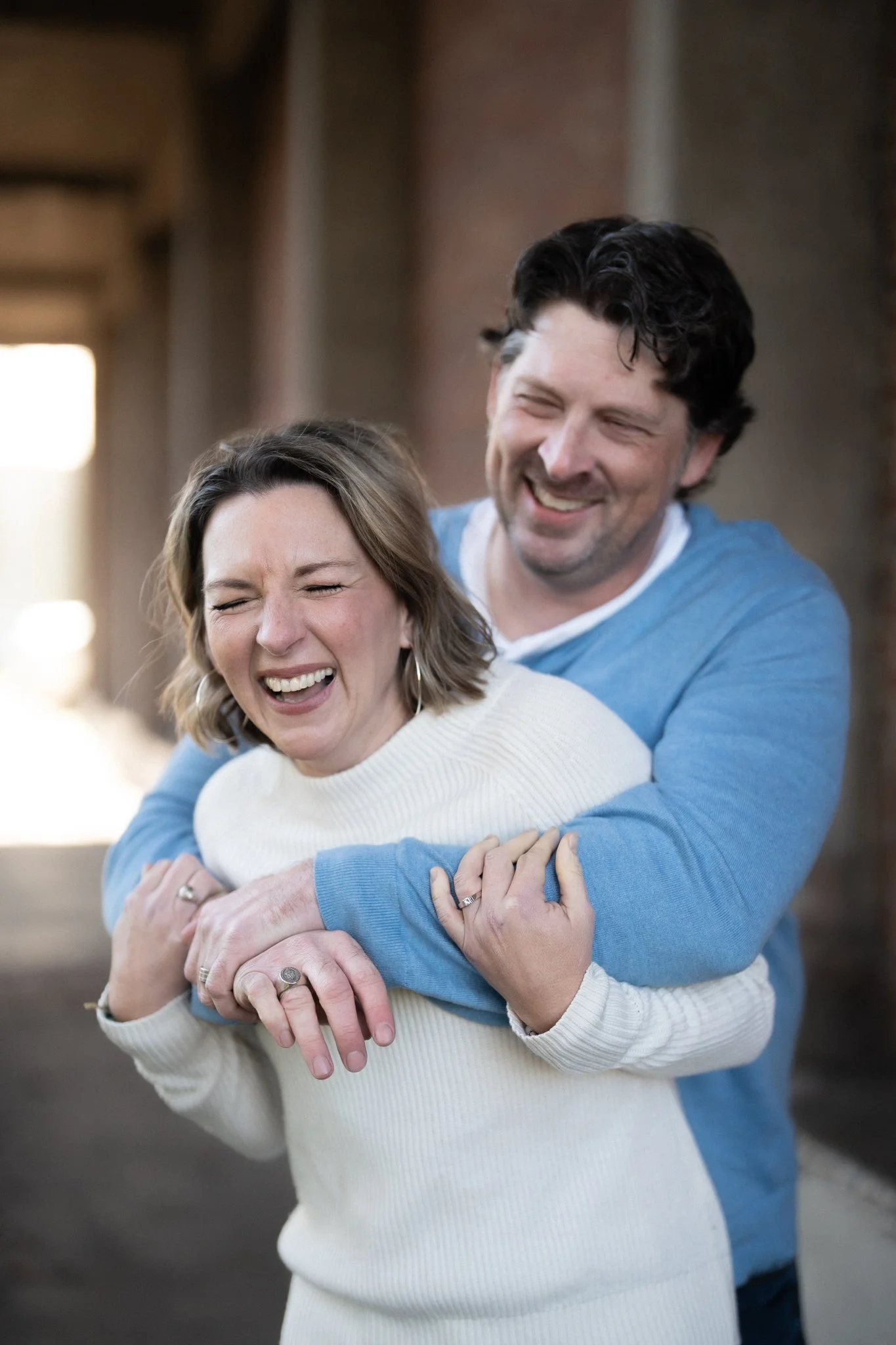 A man and woman sharing a joyful embrace indoors, smiling and laughing with their eyes closed.