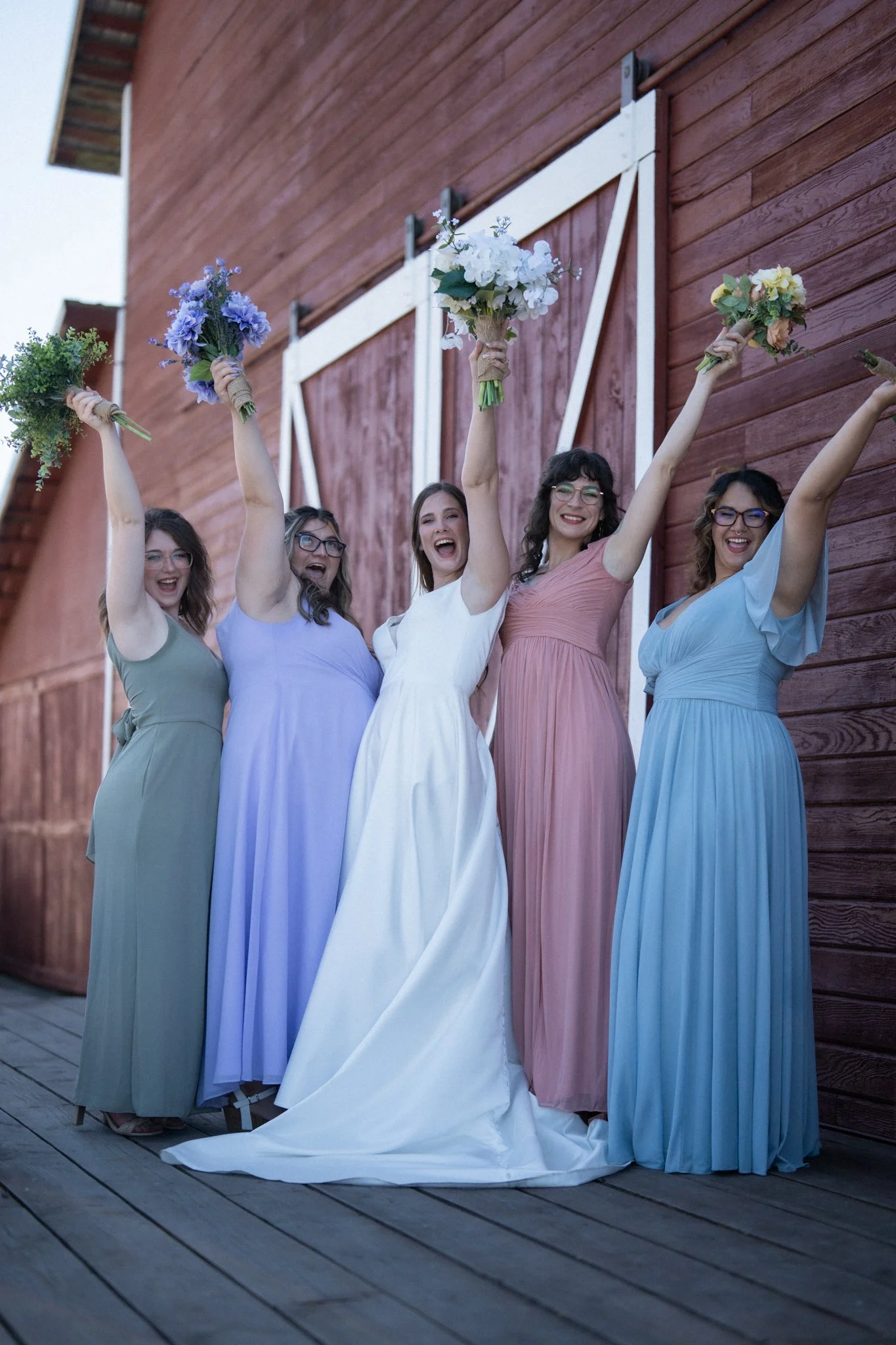Five women in pastel dresses celebrating with bouquets of flowers outside a rustic barn.