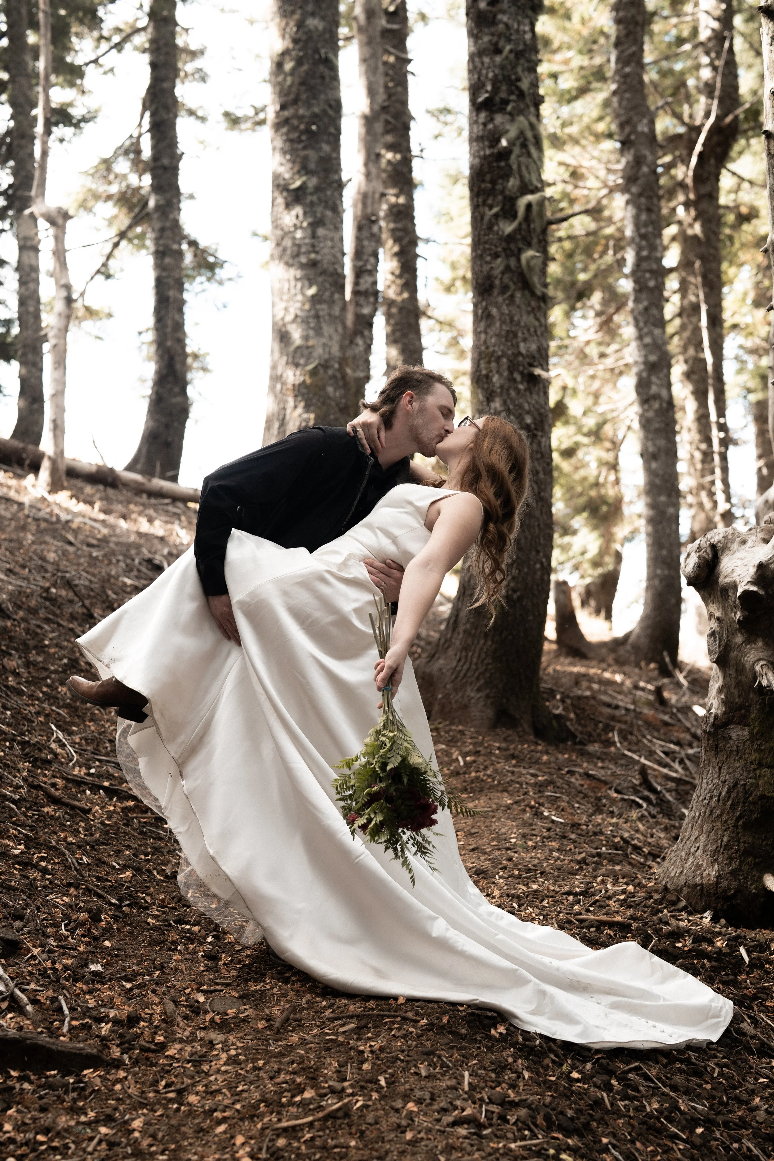 A couple sharing a kiss in a forest, with the man dipping the woman who is holding a bouquet of flowers.
