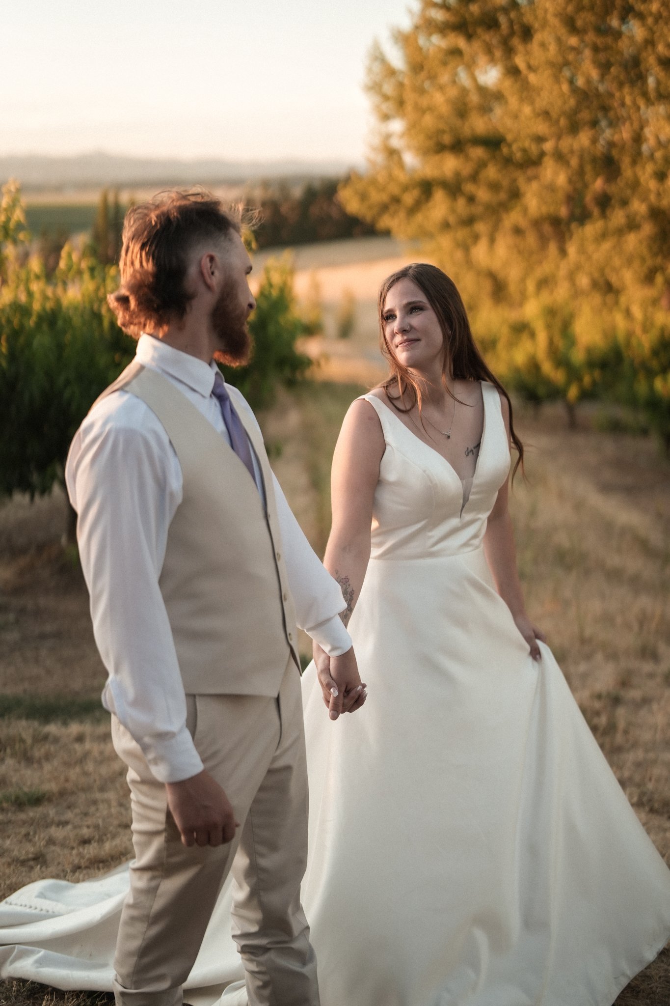 A couple in wedding attire holding hands in a vineyard at sunset.