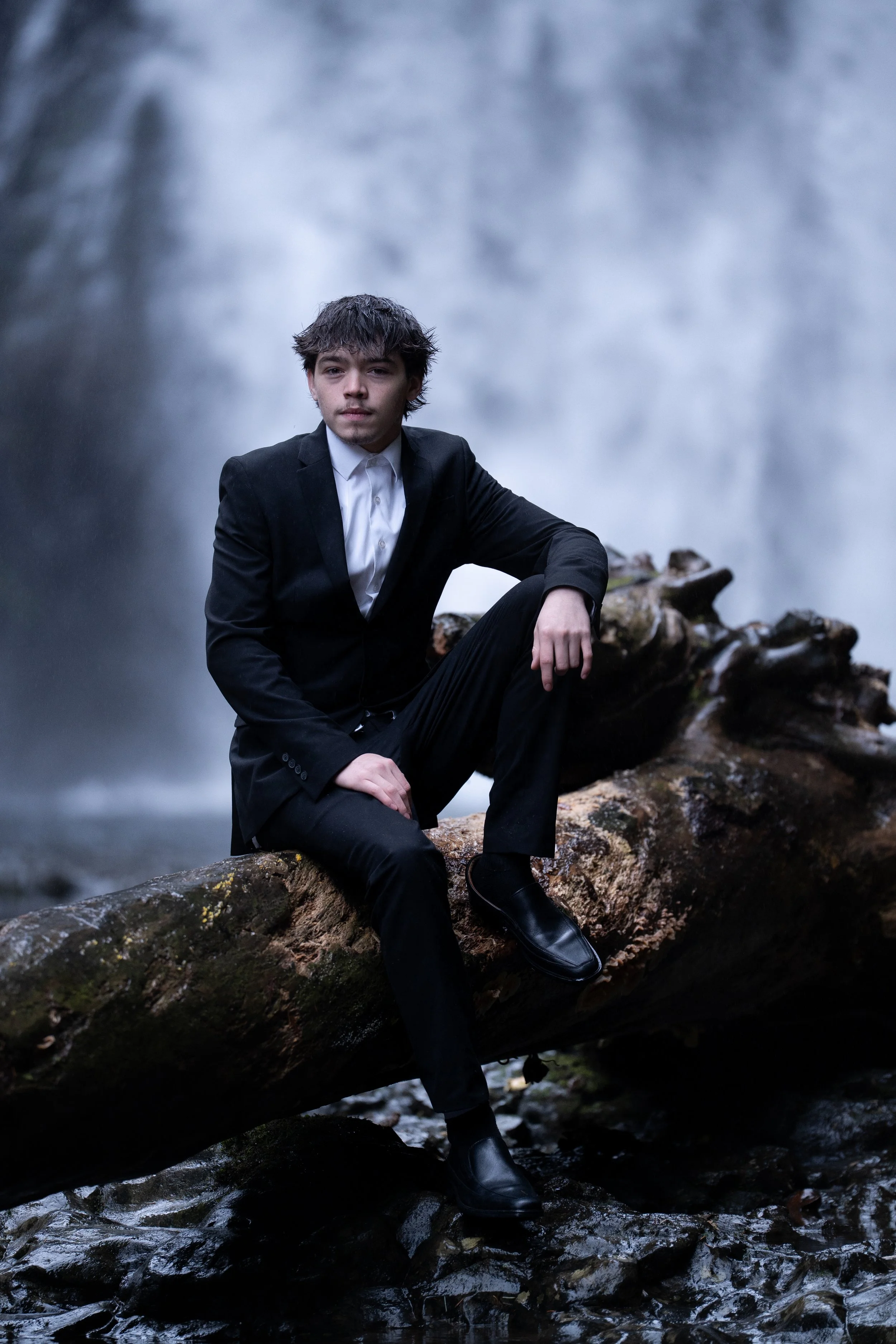 A young man in a black suit and white shirt sitting on a log in front of a waterfall.