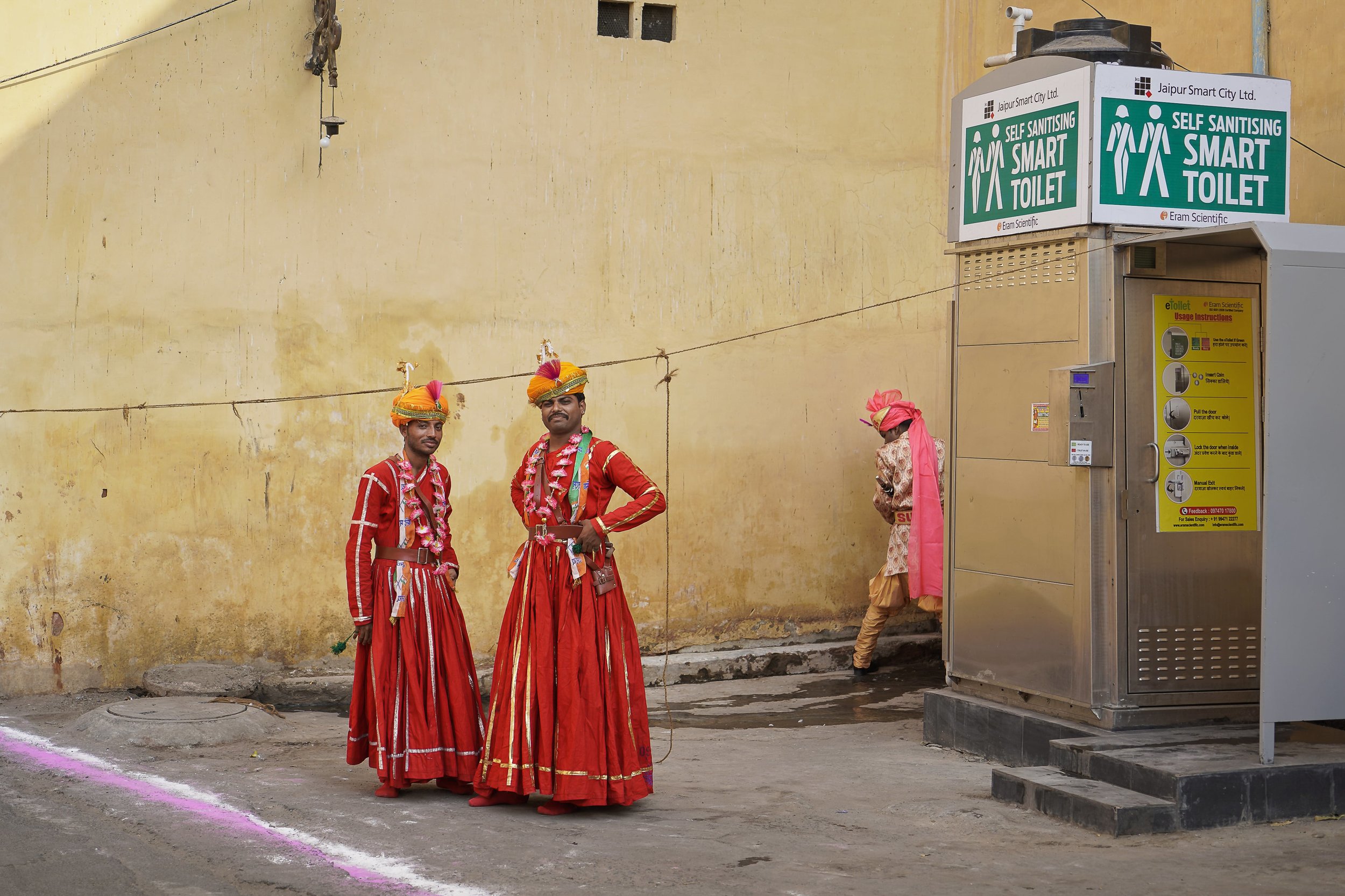 Men in traditional indian clothes with man in background urinating against wall