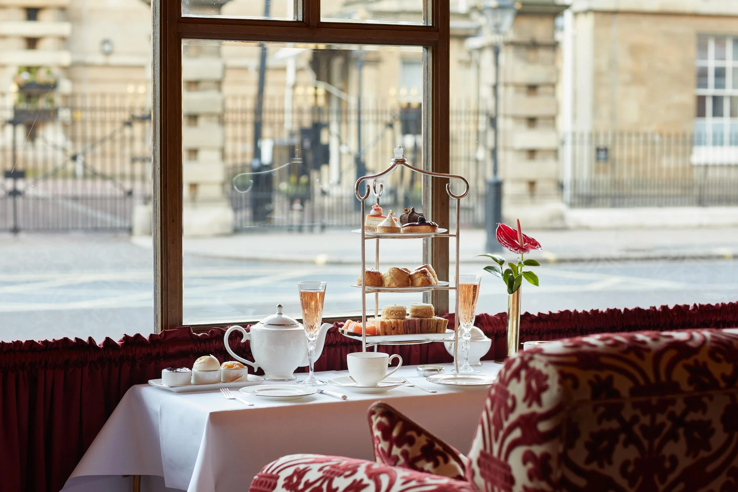 A table with an afternoon tea cake stand, tea pot, champagne glass and tea cup in front of a large window with a street view