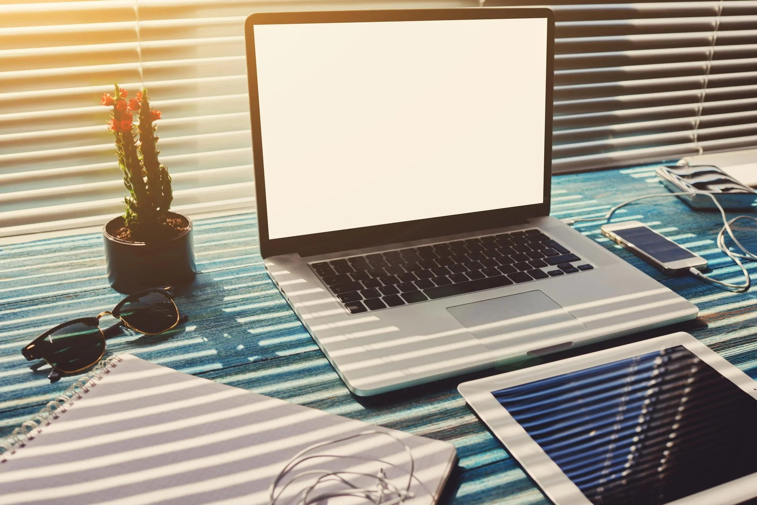 A open laptop at a desk with a window with closed blinds behind it, a plant, some paper, a tablet and headphone on the desk with it