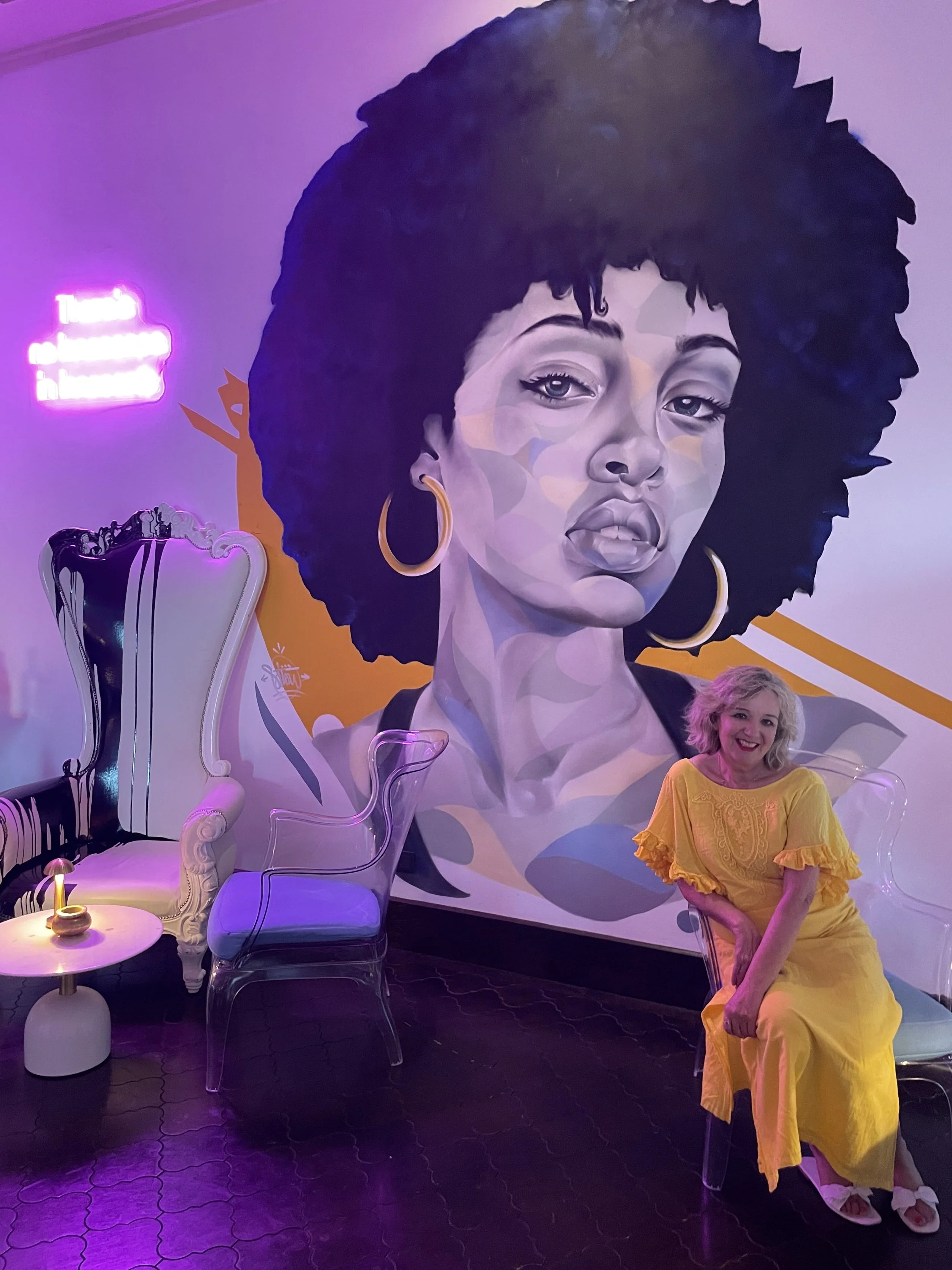 Wendy Rigg wearing a yellow dress sitting in from of a large portrait of a woman with an afro