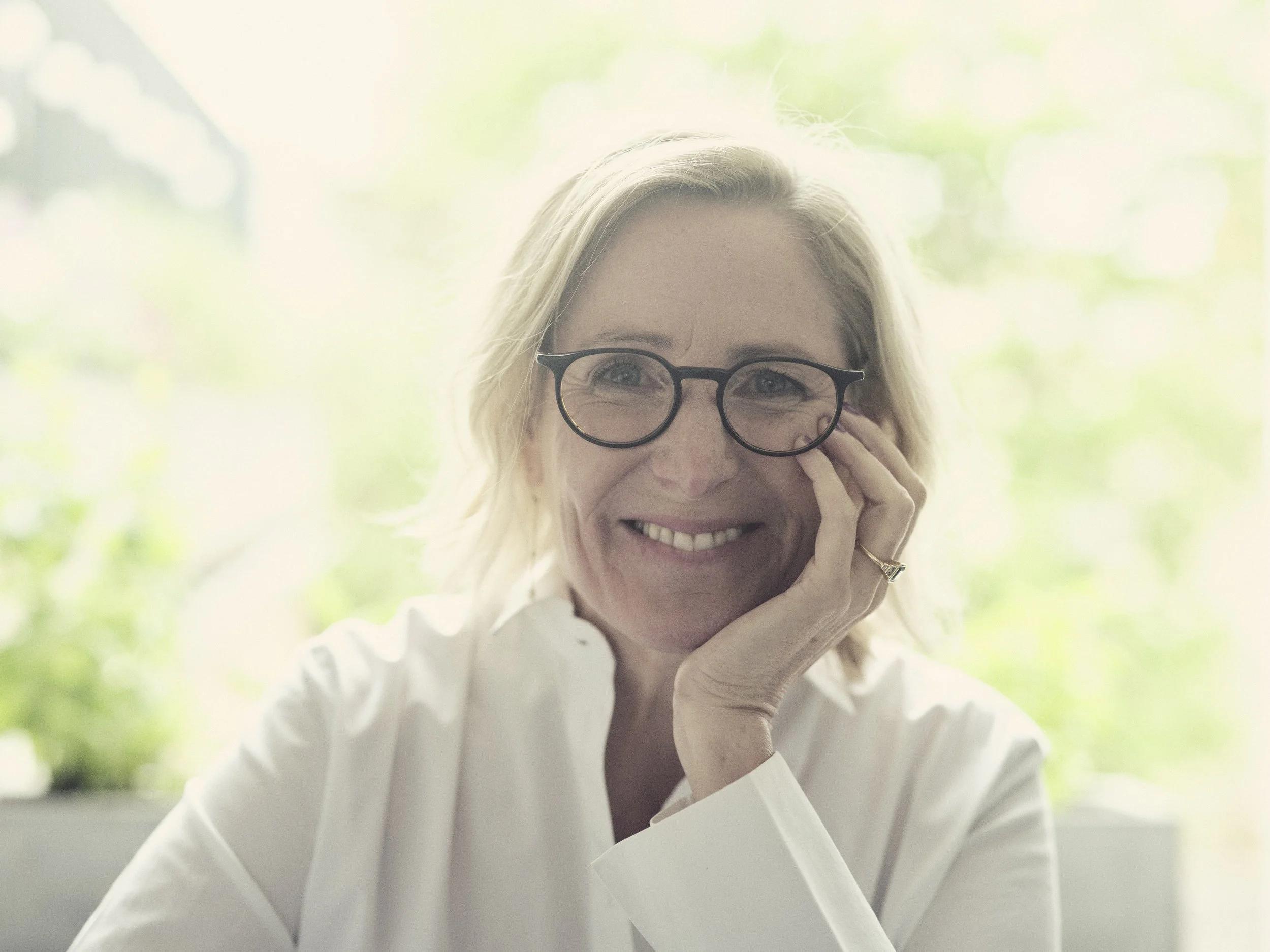 Anna Fielding with blonde hair and waring black framed glasses smiling at the camera as she hold her left hand to her face