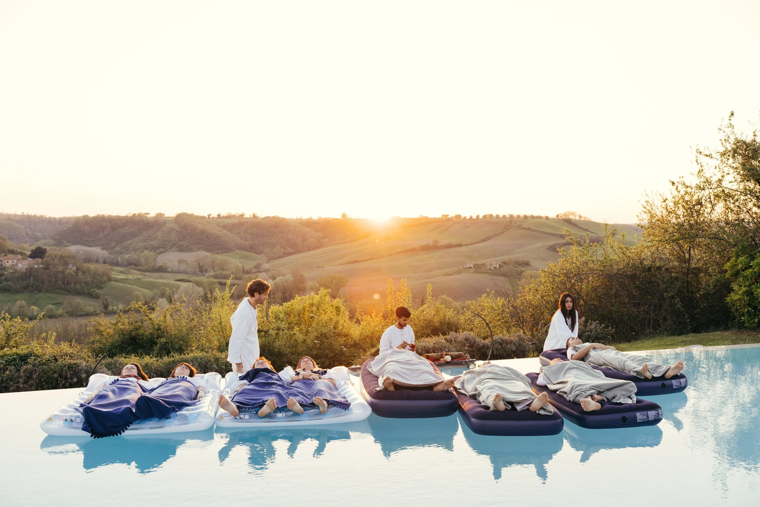 A group of people lying on inflatable beds floating in a pool with sunset and mountains in the background