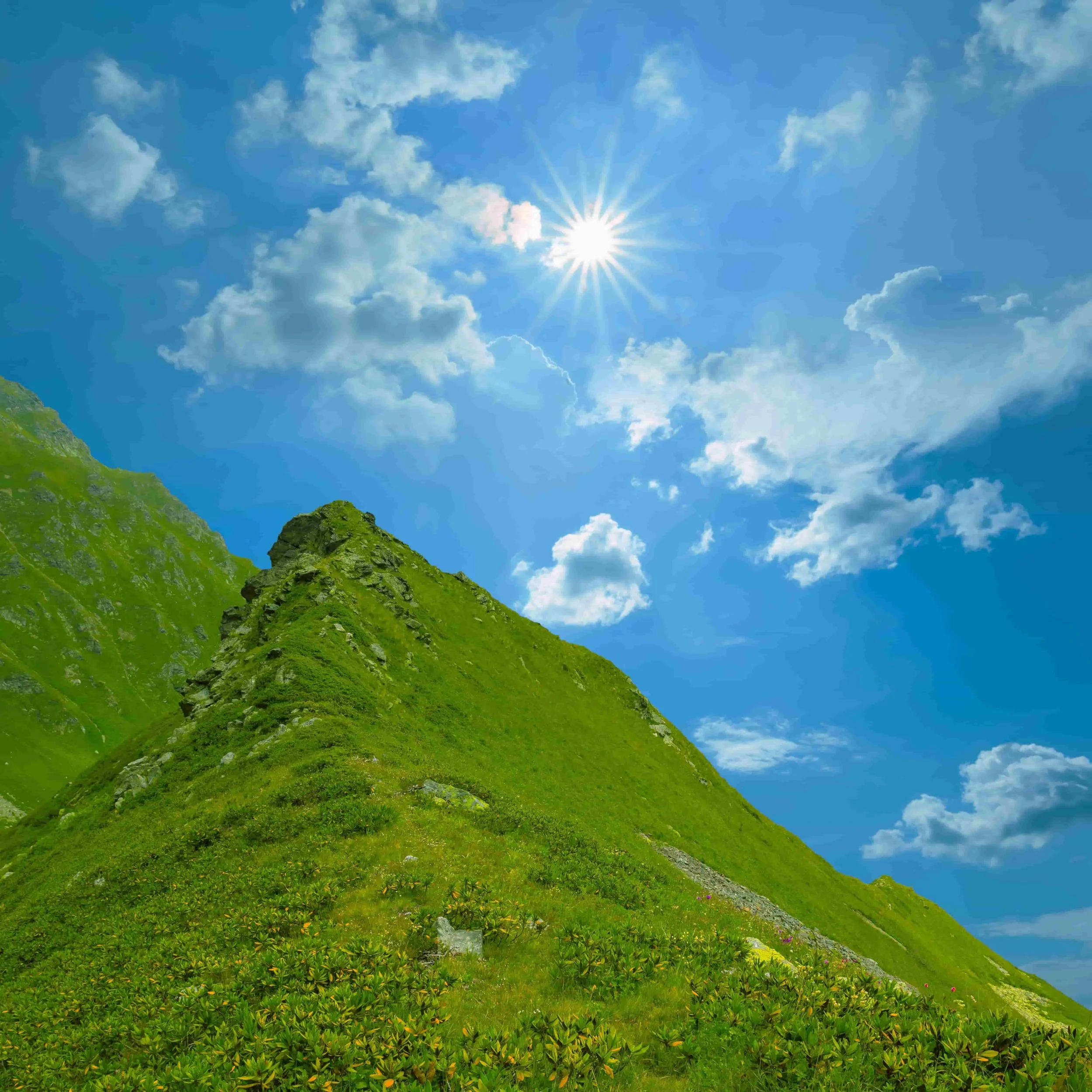 Bright sun in blue sky with scattered clouds over green mountain with grassy slope.