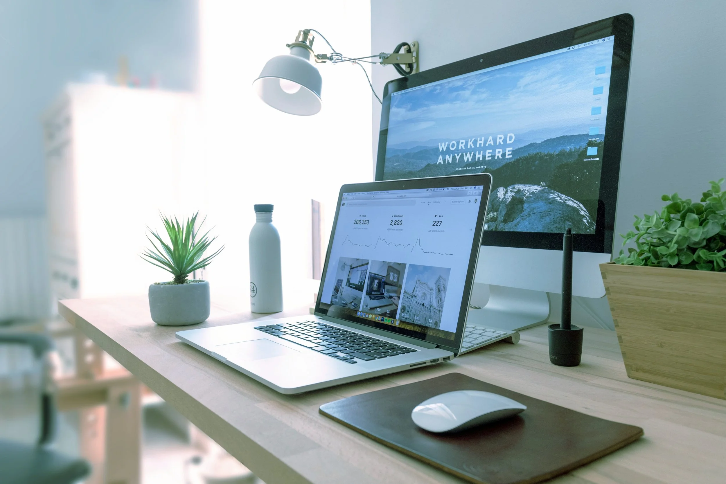 A Macbook laptop placed in front of a computer screen on a desk with a plant, water bottle, lamp and computer mouse surrounding them