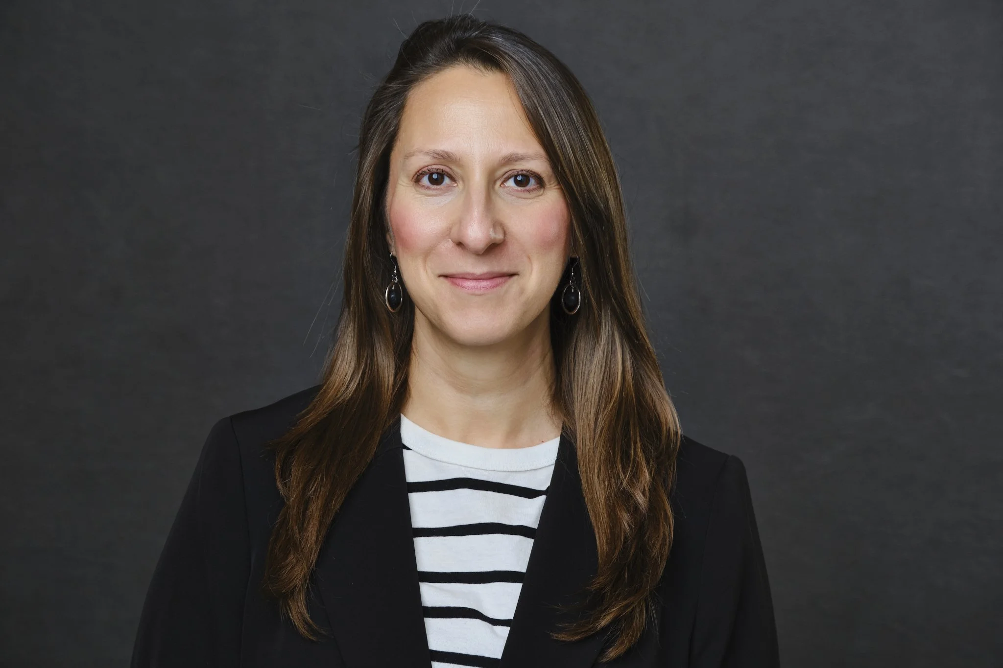 A woman with long brown hair, stands in front of a gray background. She is wearing a black and white stripped blouse under a black blazer. She is smiling at the camera.
