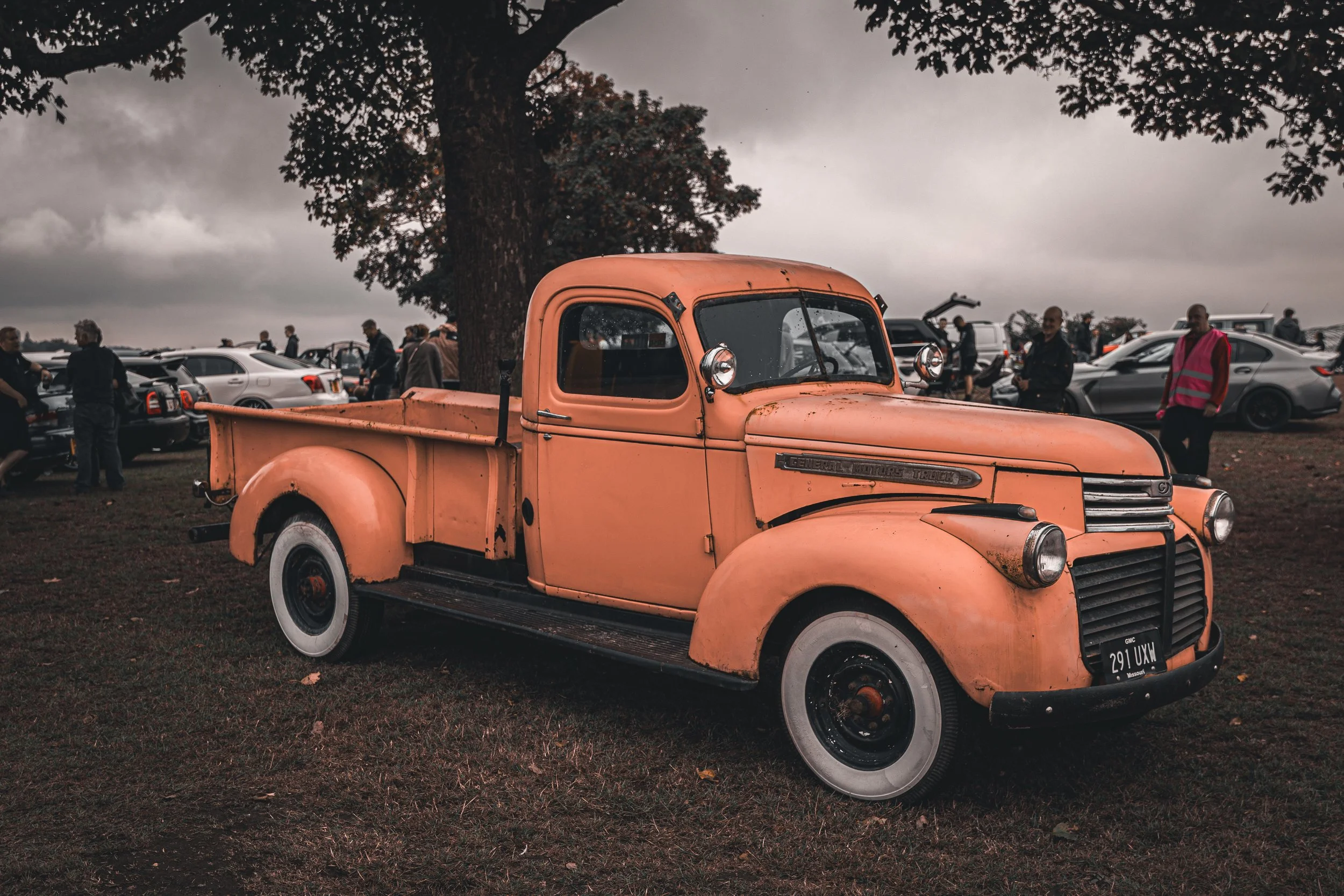 Vintage orange truck parked outdoors at a car show with other cars and people in the background under an overcast sky.