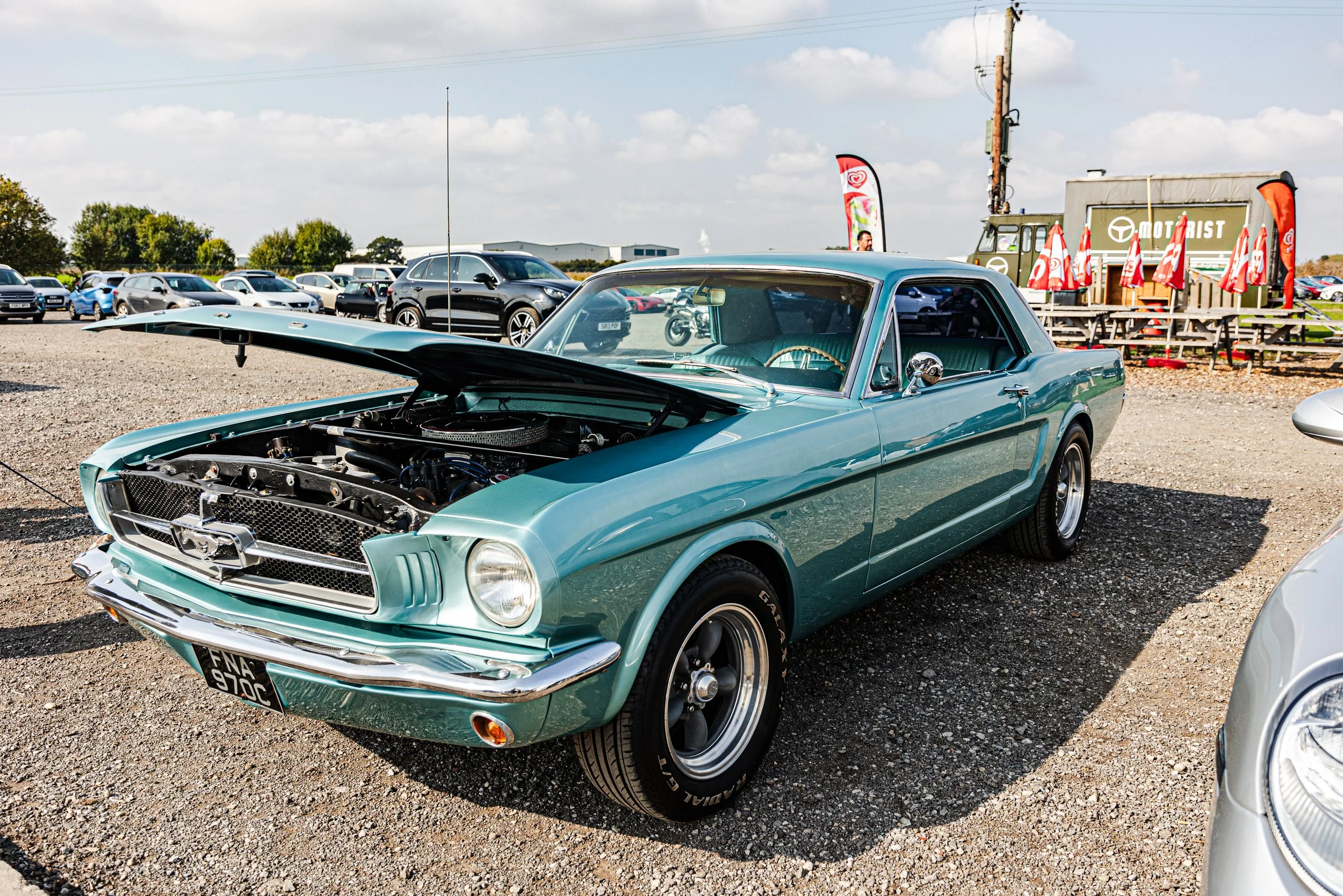 A mint green vintage Ford Mustang car on display at a car show, with its hood open revealing the engine. The background shows other cars, a small outdoor seating area with red umbrellas, and flags on display.