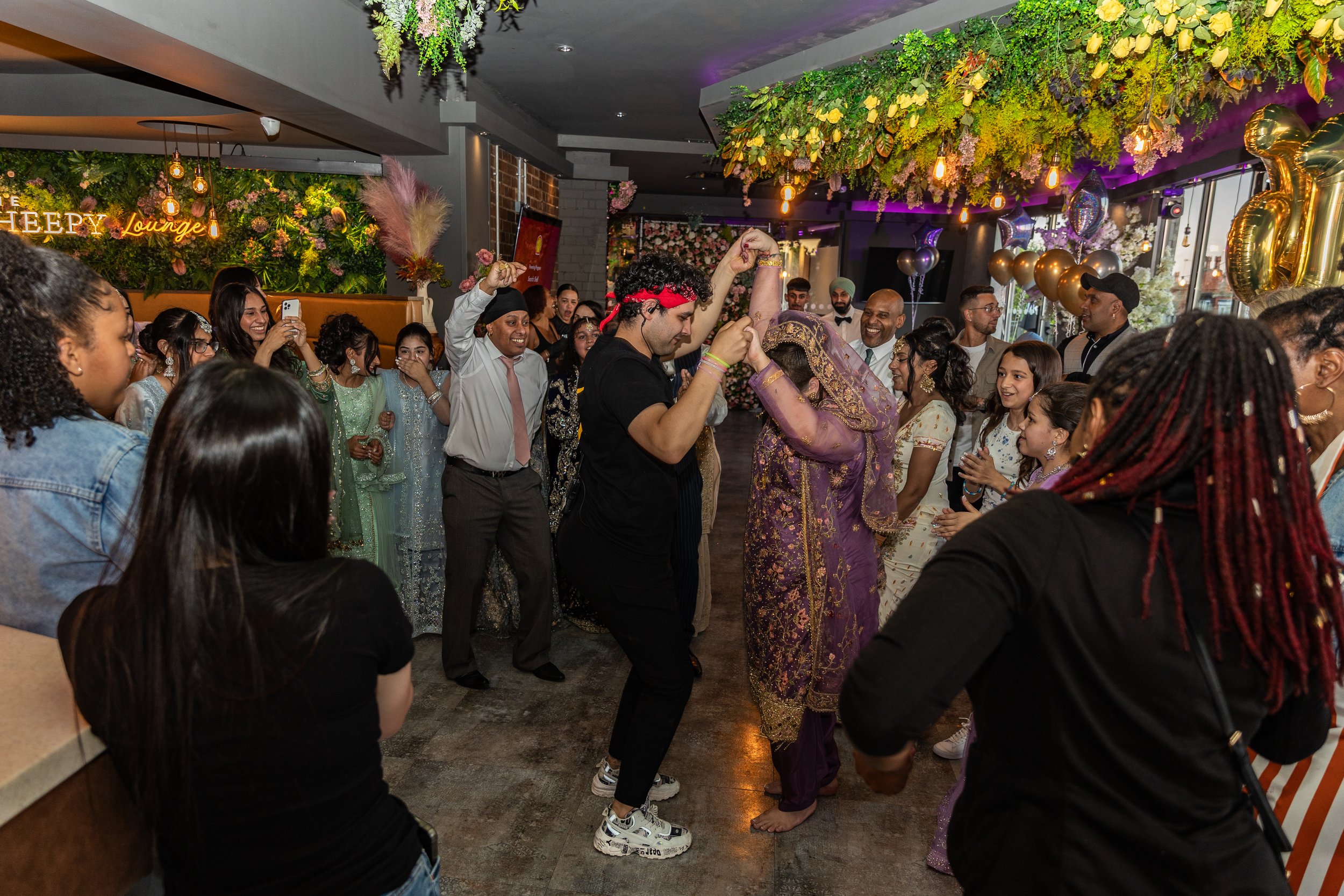 People dancing and celebrating at a party in a decorated indoor venue with floral and balloon decorations, including a sign that says 'Chery Lounge' in the background.