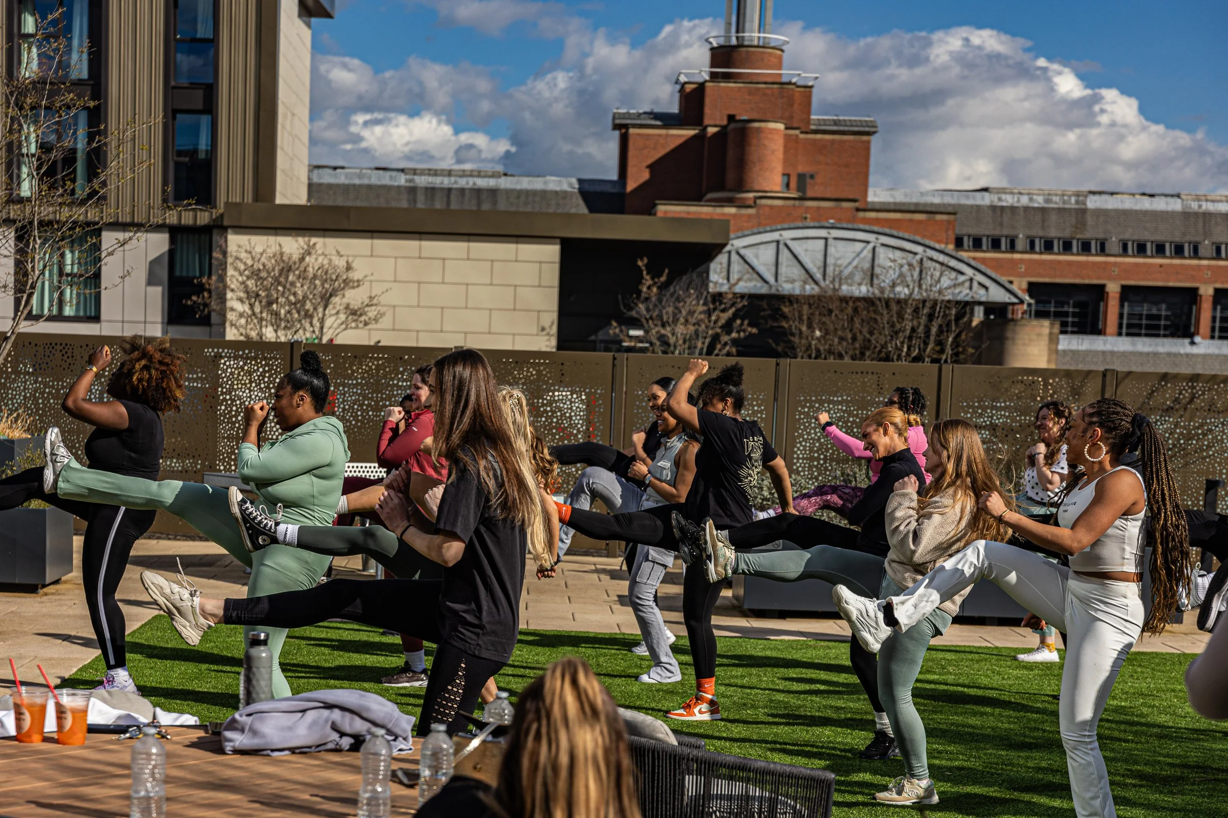 A group of women participating in an outdoor fitness class, performing high-knee kicks on a grassy area with a cityscape in the background.