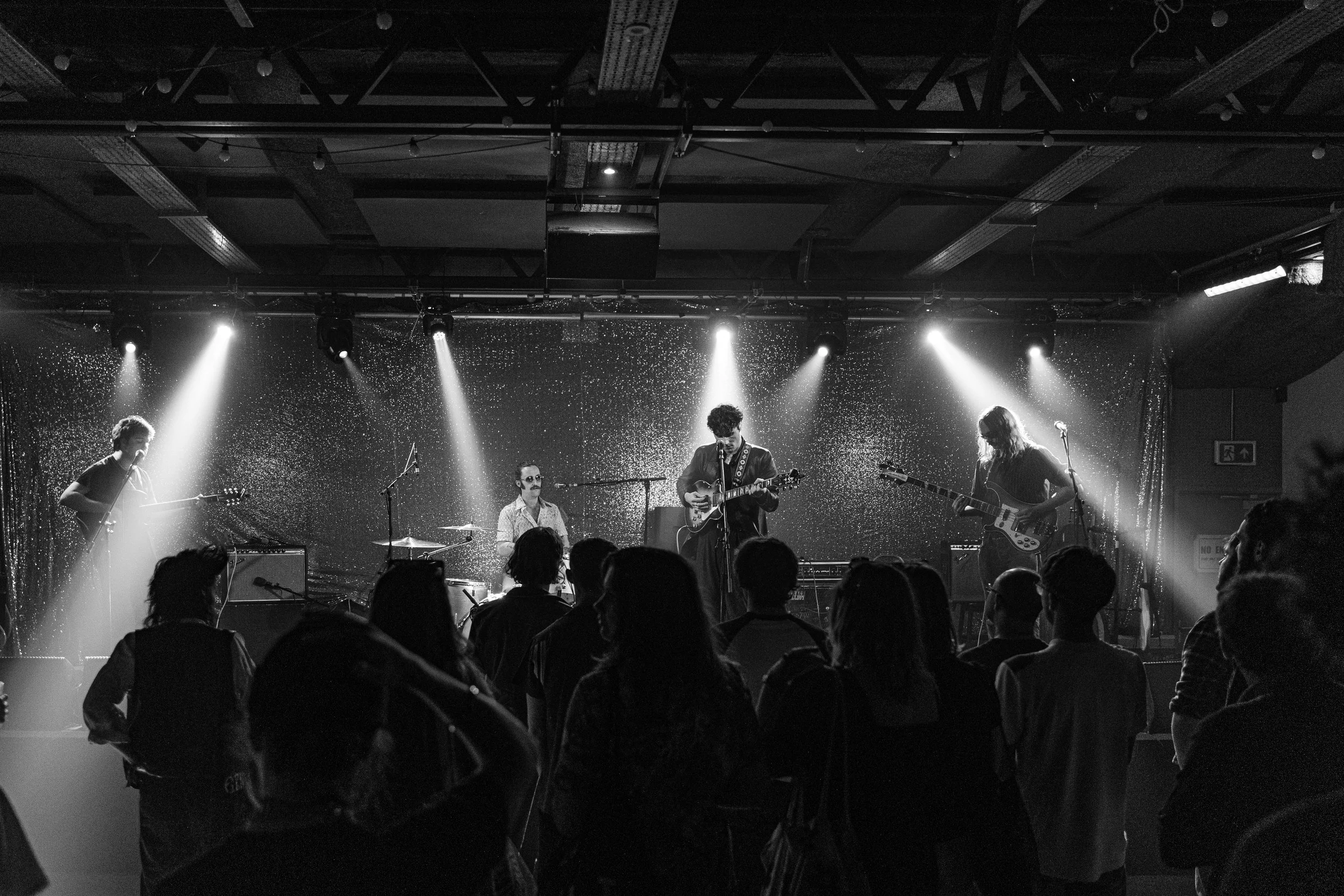 Black and white photo of a live band performing on stage with four musicians playing guitars and drums, and an audience watching in front of the stage.