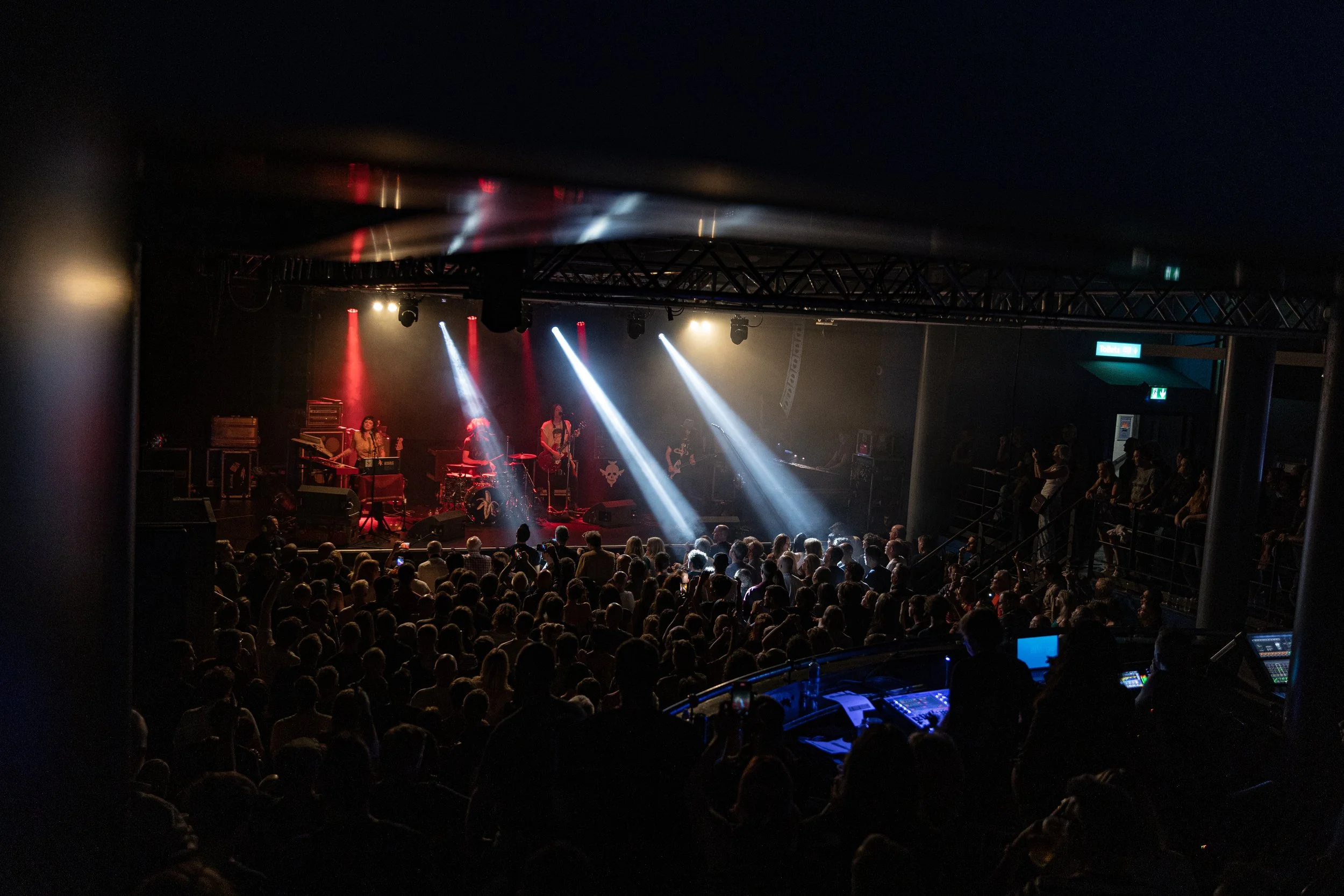 Concert scene with band performing on stage in front of an audience, stage lighting with red, white, and blue beams, and audience members watching from a darkened venue.