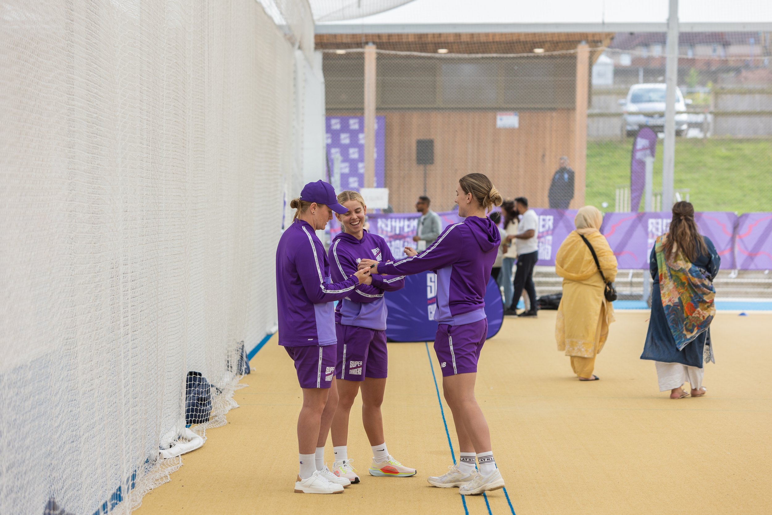 Three women in purple athletic outfits standing on a yellow sports court, interacting and smiling, with two women in traditional clothing in the background. The scene appears to be at an outdoor sports or event venue.