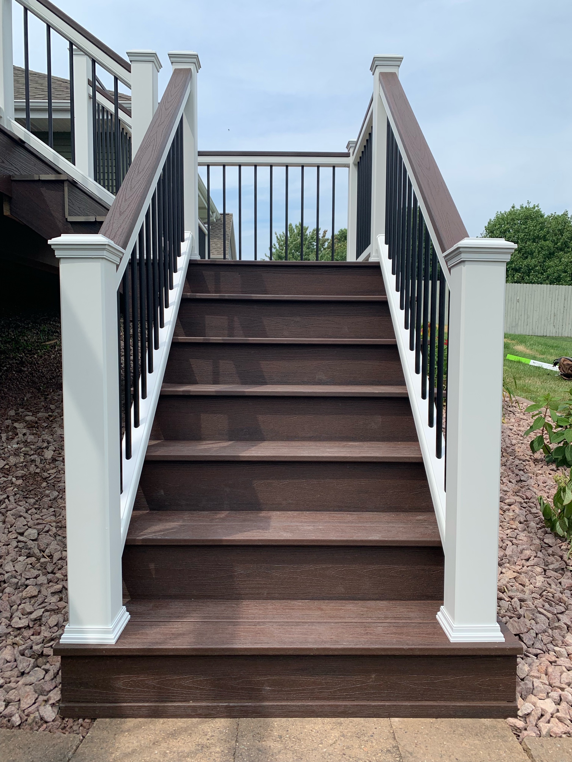 Trex Lava Rock deck boards with white top and bottom rails, Lava Rock drink rail on top of railings, black aluminum balusters and white post sleeves, post skirts, post caps. Double picture frame around perimeter of deck.