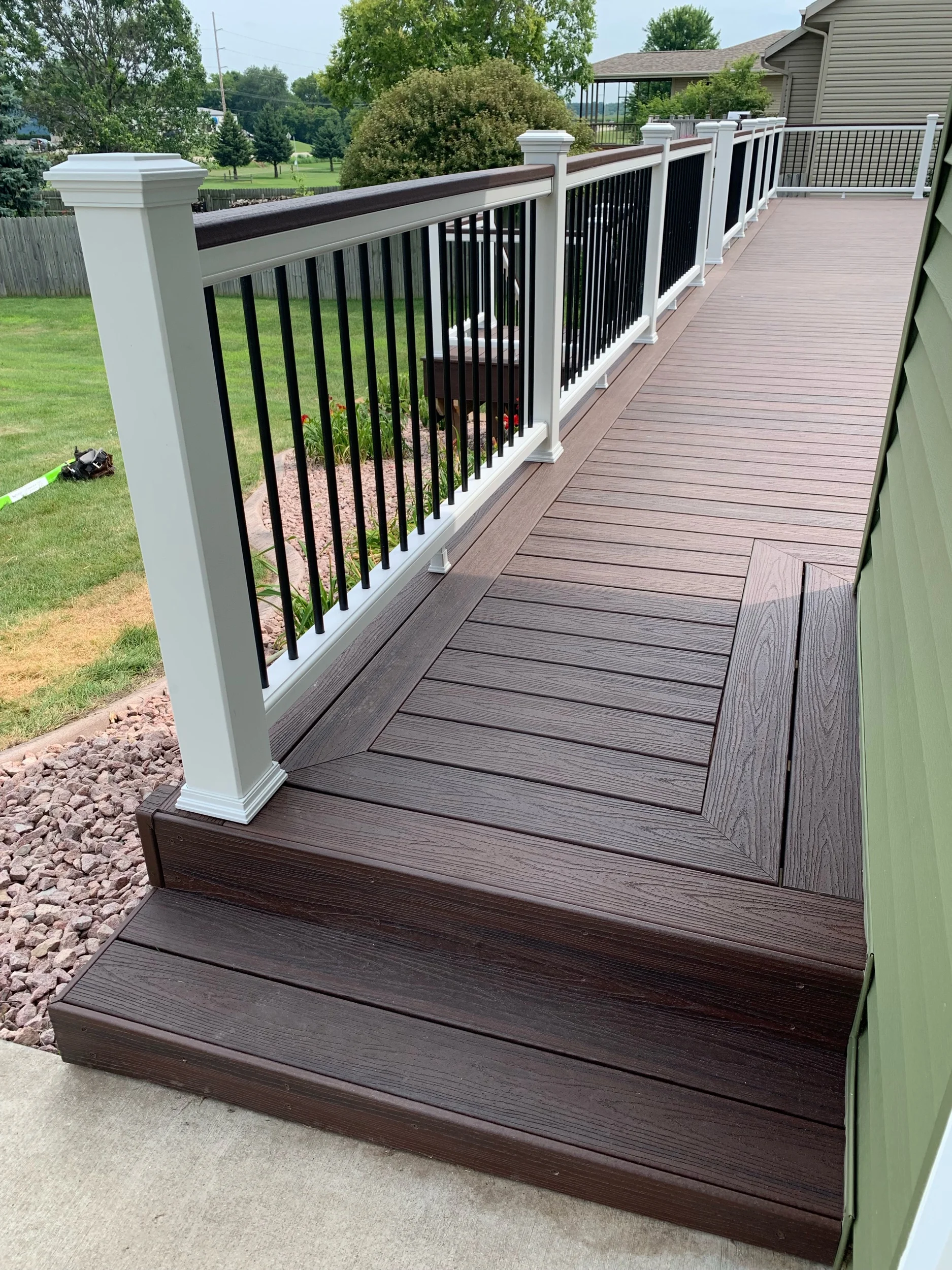 Trex Lava Rock deck boards with white top and bottom rails, Lava Rock drink rail on top of railings, black aluminum balusters and white post sleeves, post skirts, post caps. Double picture frame around perimeter of deck.