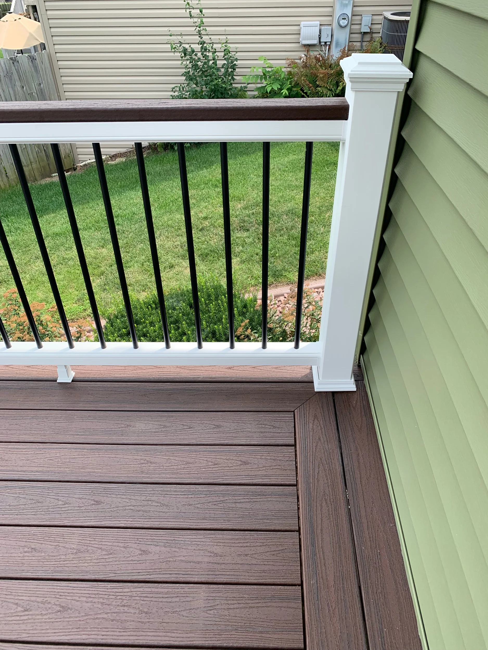 Trex Lava Rock deck boards with white top and bottom rails, Lava Rock drink rail on top of railings, black aluminum balusters and white post sleeves, post skirts, post caps. Double picture frame around perimeter of deck. 