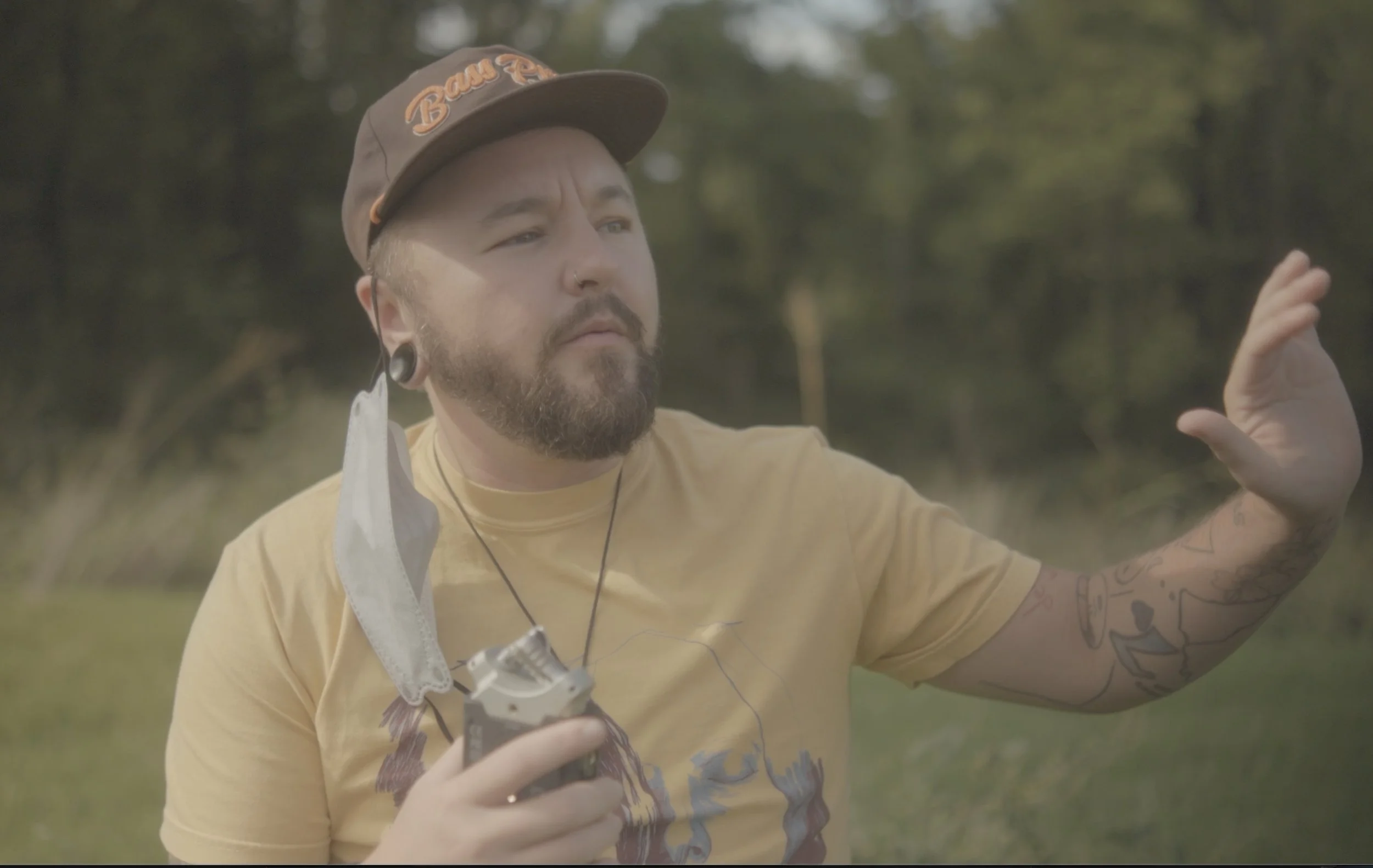 basil, a man, in a yellow shirt and brown hat, holding Zoom recorder, in a field in Memphis, TN