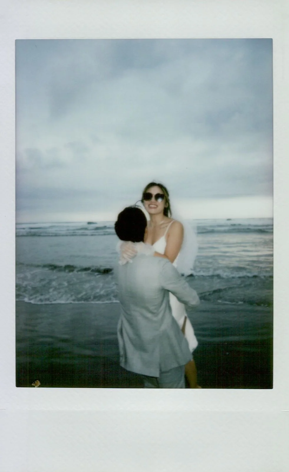Polaroid photo of bride in white silk dress and groom in light suit walking barefoot on Cannon Beach, Haystack Rock in the distance