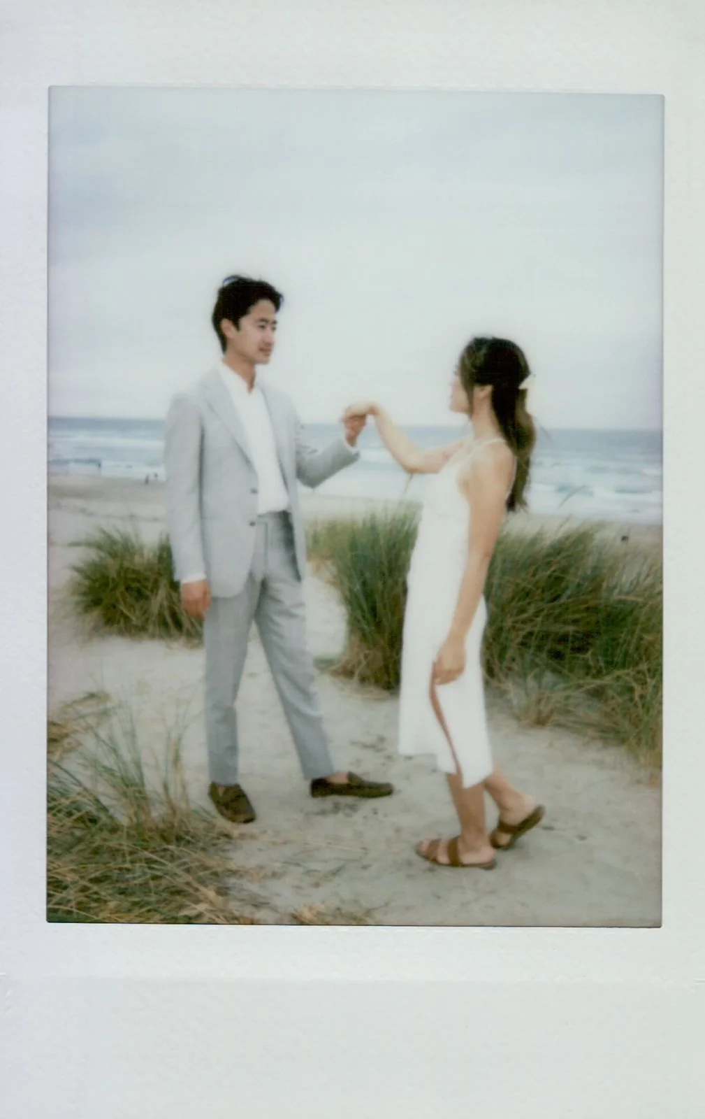 Polaroid photo of bride in white silk dress and groom in light suit walking barefoot on Cannon Beach, Haystack Rock in the distance