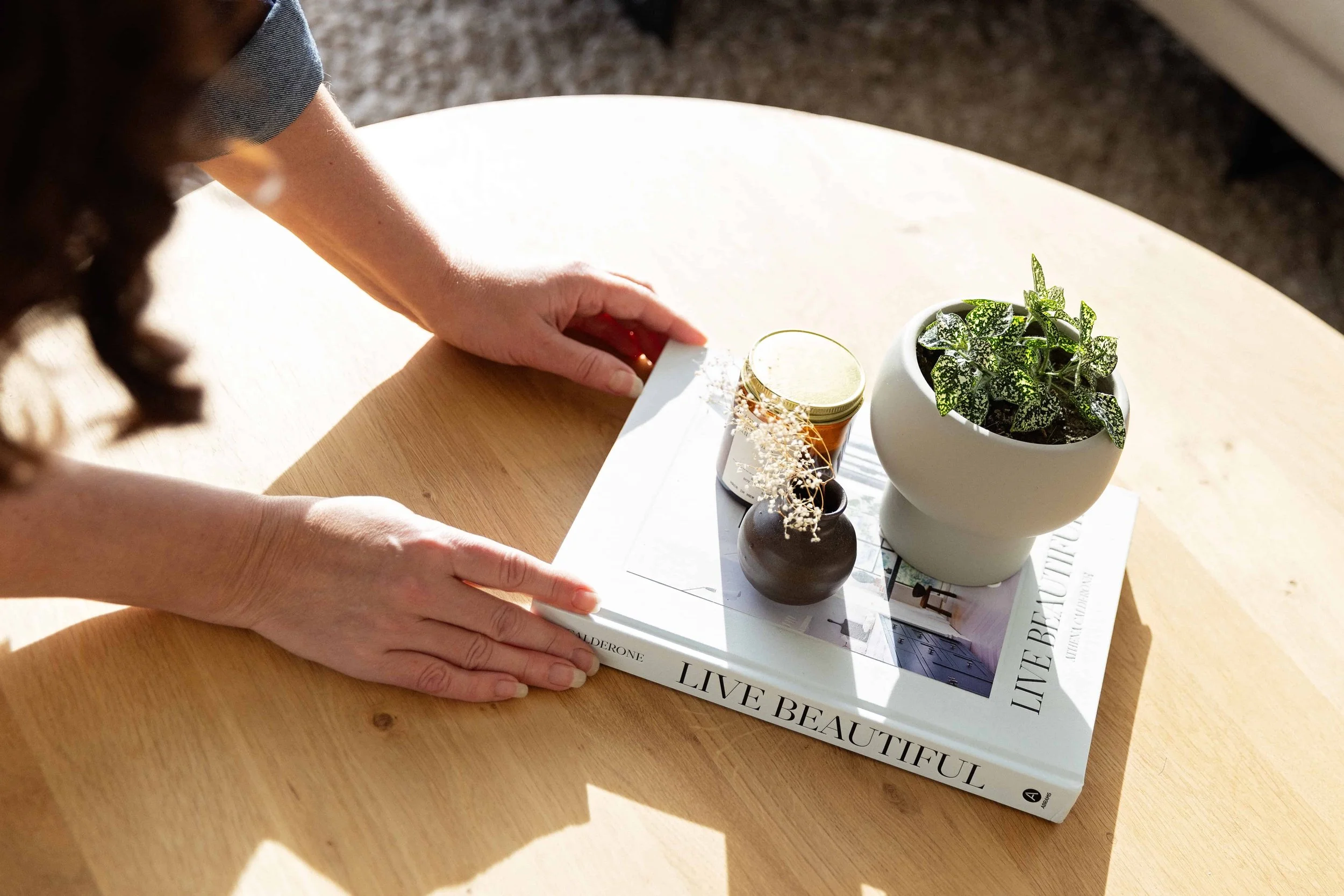 A person arranging a tray with a potted plant, a small jar, and a mini vase on top of a large book on a wooden table.