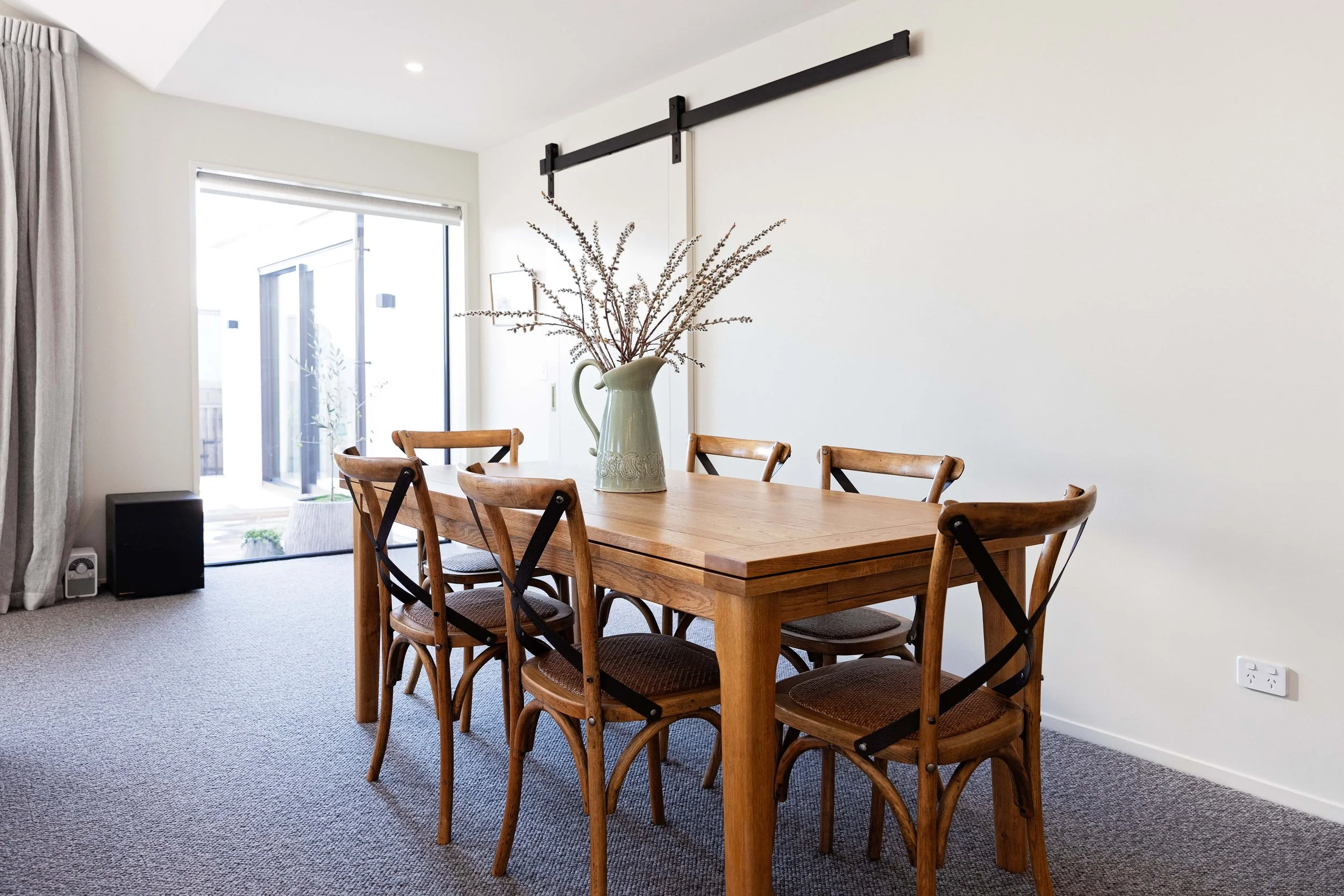 Dining room with a wooden table, six matching chairs, a large vase with dried flowers, sliding glass doors, beige curtains, and a carpeted floor.
