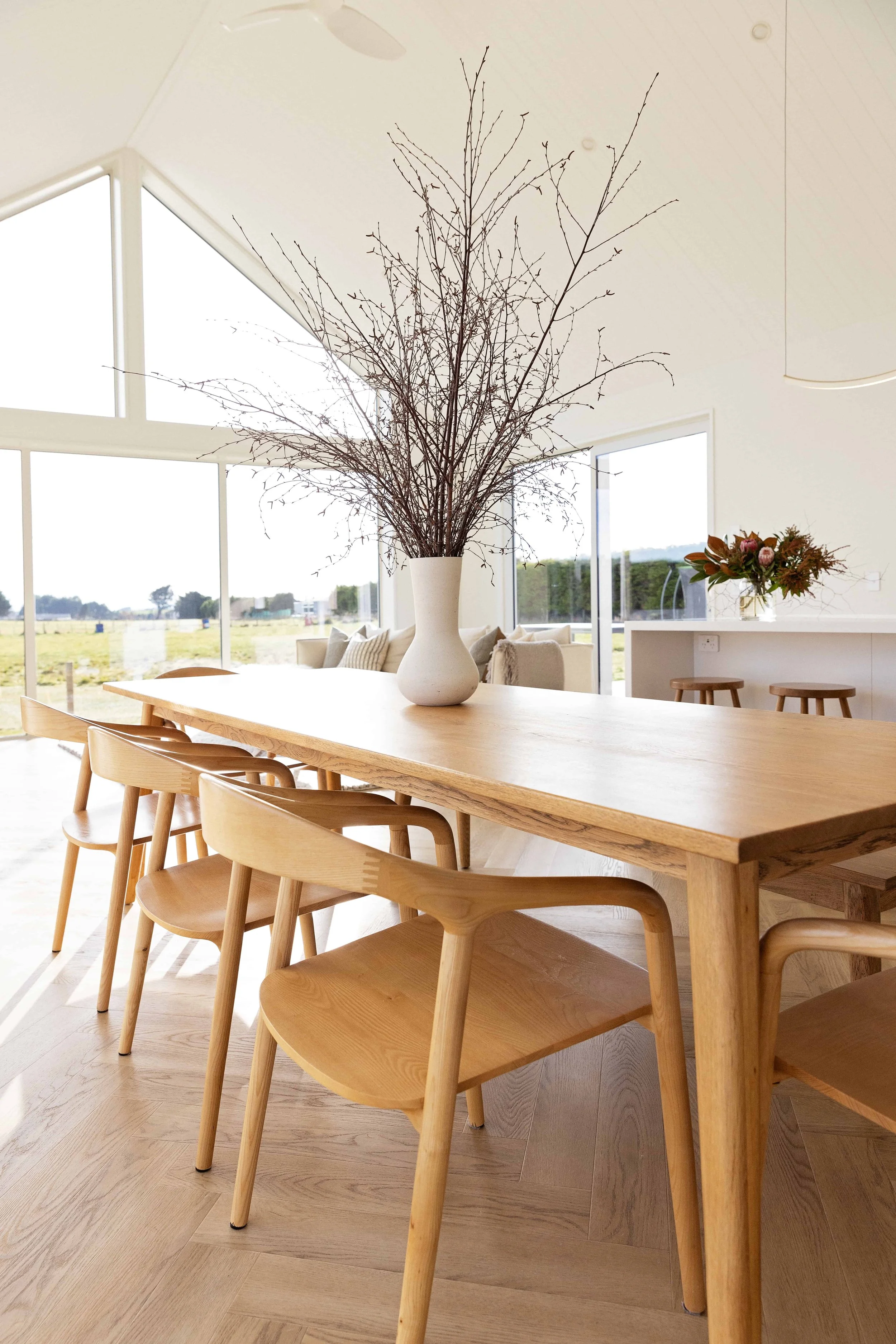 Interior of a bright, modern dining area with a long wooden table, natural wood chairs, a large white vase with bare branches, and a window view of an outdoor field.