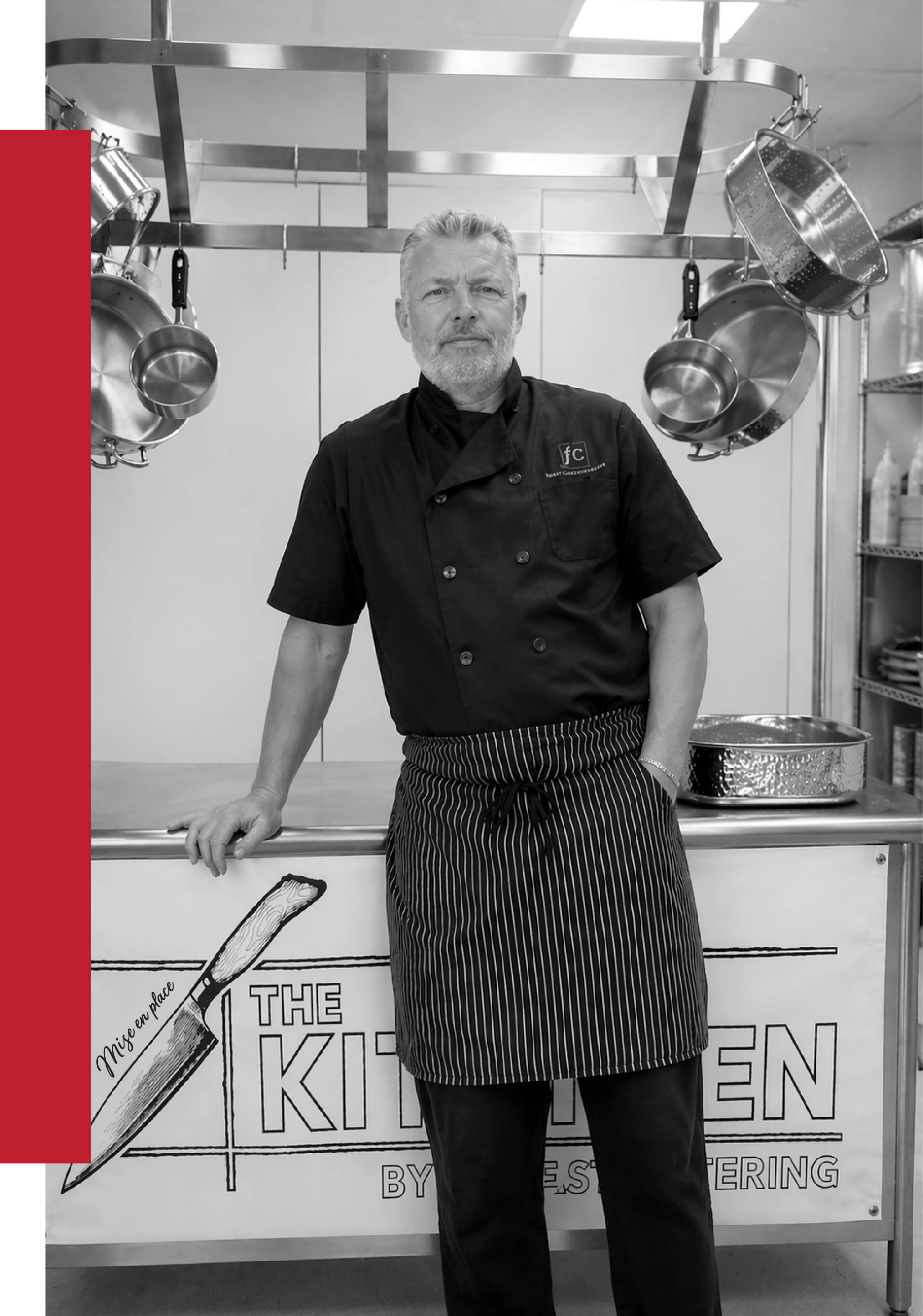 A male chef with gray hair and a beard, wearing a black chef's coat and striped apron, standing in a commercial kitchen with hanging stainless steel pots and pans in the background.