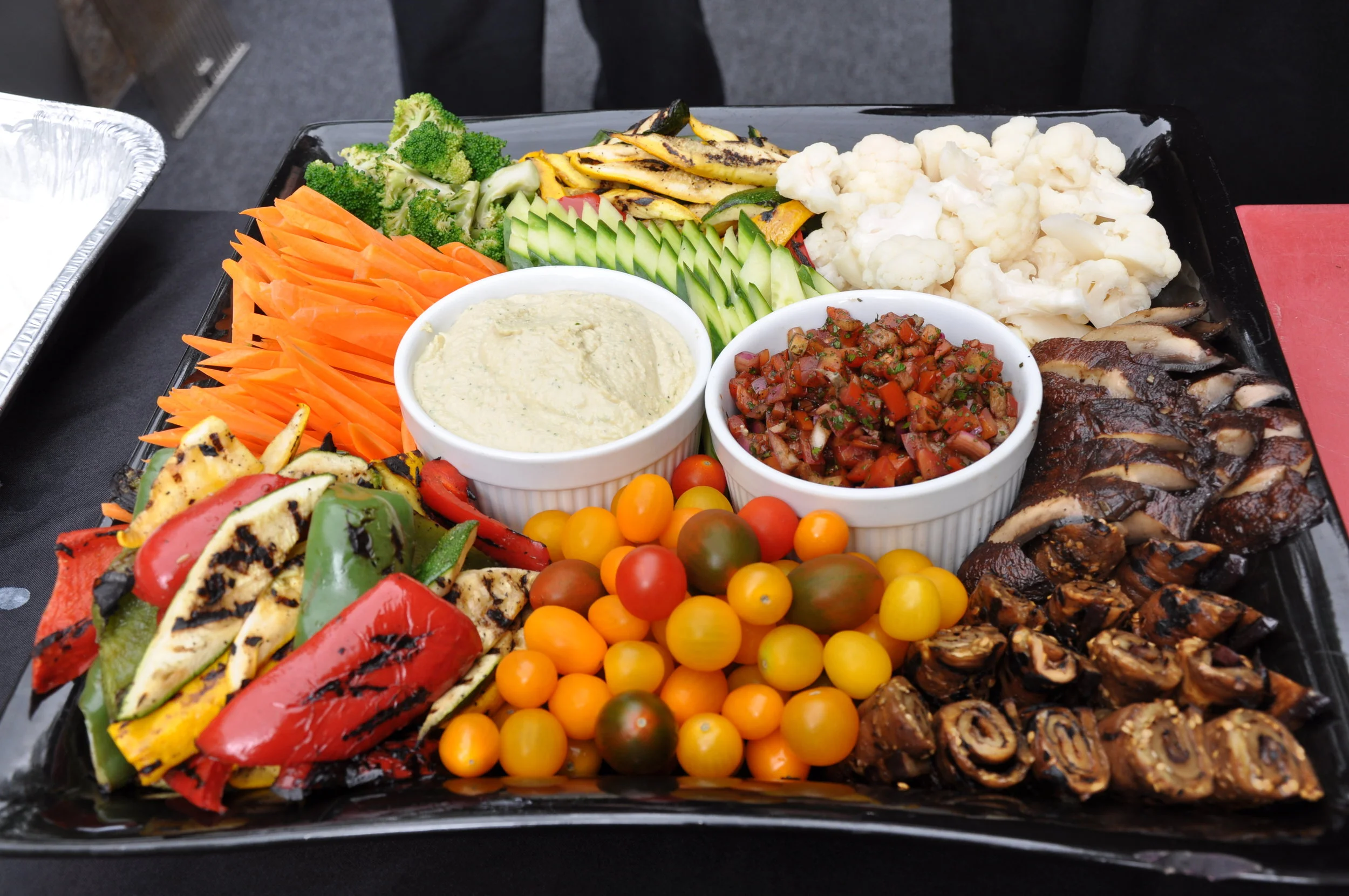 A platter of grilled vegetables and assorted fresh vegetables including cherry tomatoes, sliced cucumbers, broccoli, cauliflower, grilled zucchini, and grilled peppers, with two bowls of dips or sauces.