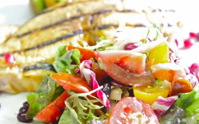 Fresh vegetable salad with tomatoes, lettuce, carrots, and olives, served with grilled fish fillets in the background.