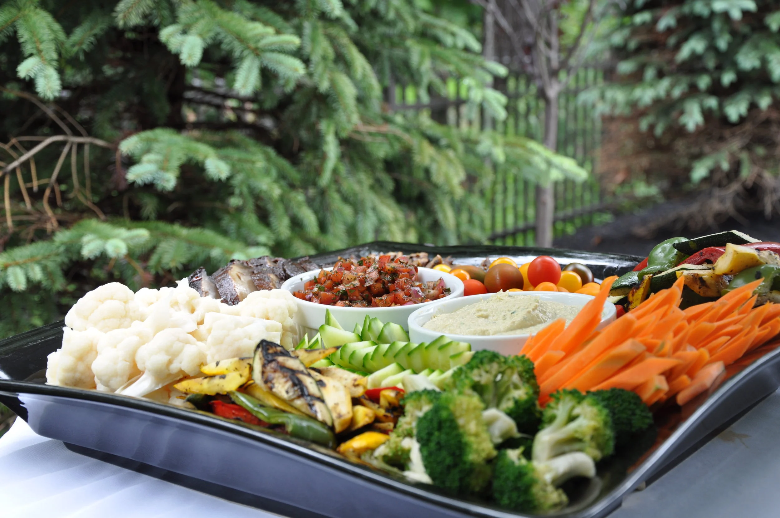 Assorted vegetables like cauliflower, cucumbers, carrots, broccoli, and grilled zucchini; dishes including salsa, hummus, and grilled meats on a black tray outdoors.