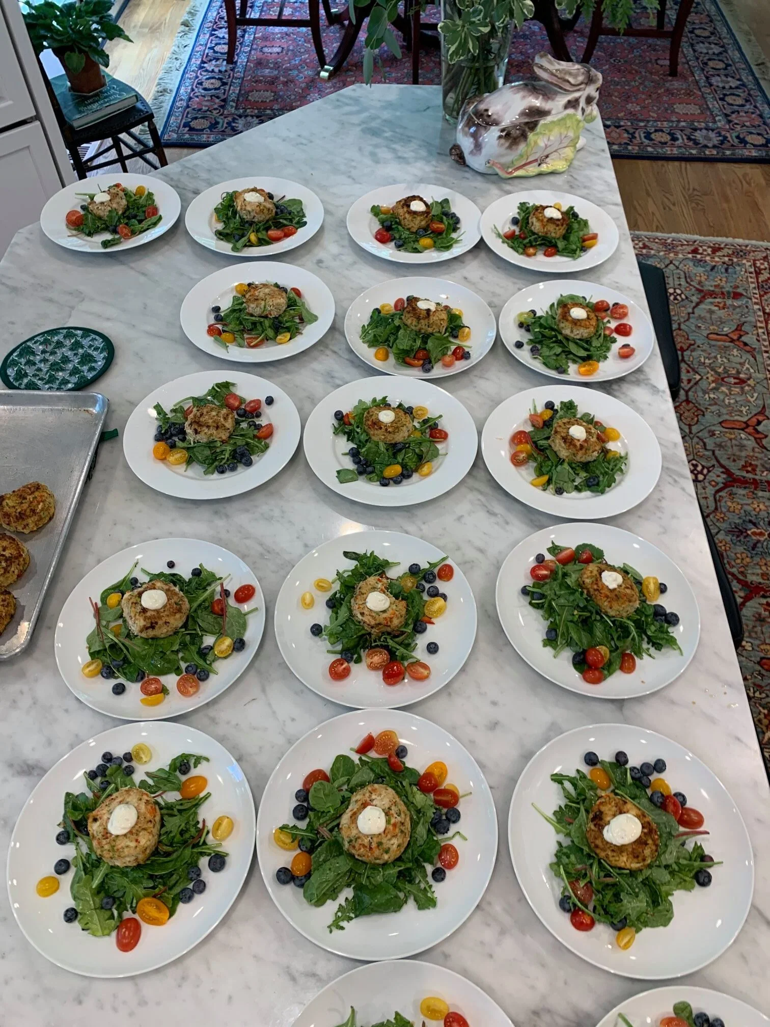 Multiple white plates with salads containing leafy greens, cherry tomatoes, blueberries, and a croquette with a dollop of sauce on top, arranged on a marble table.