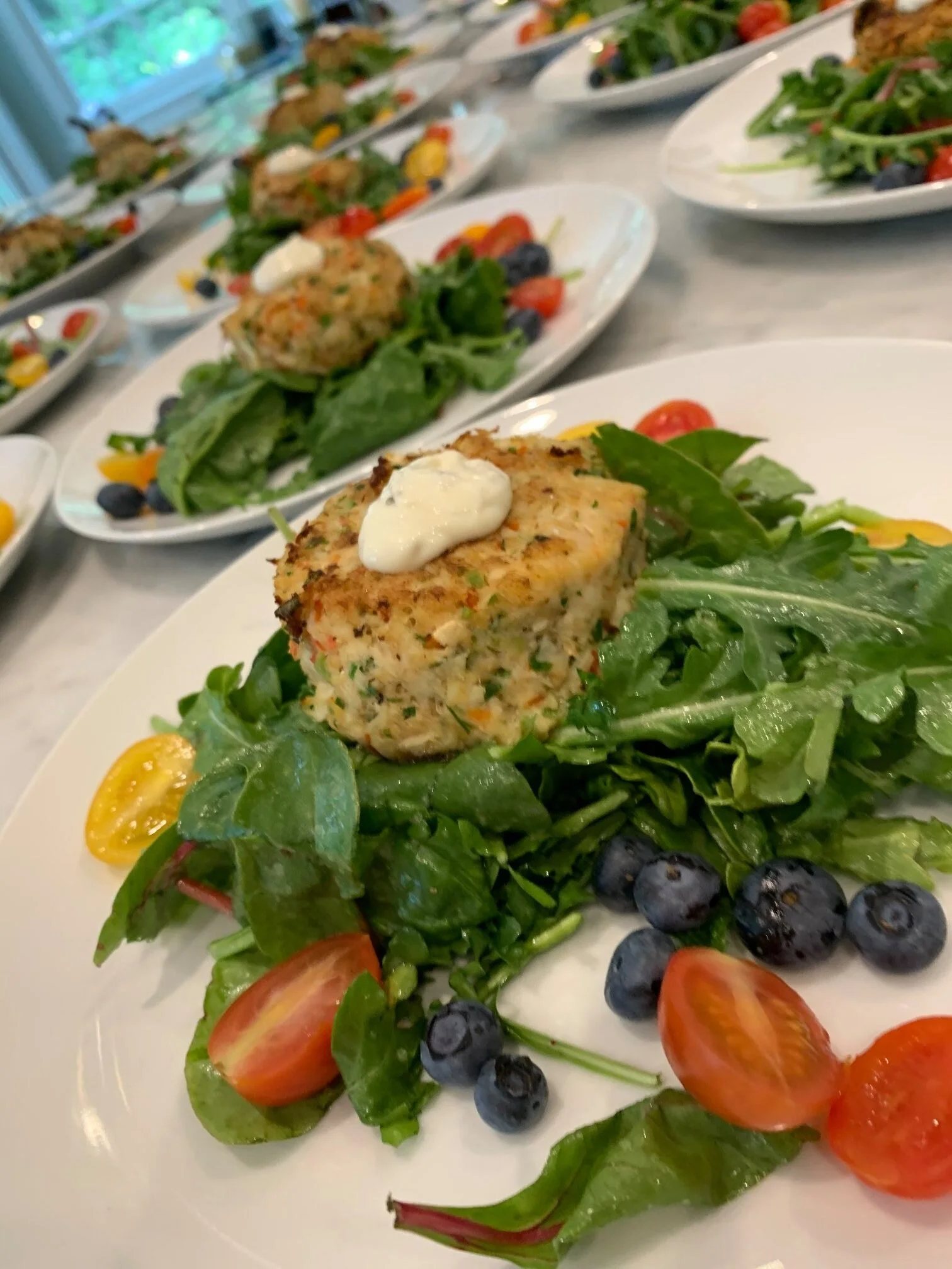 Multiple white plates with salads featuring leafy greens, cherry tomatoes, blueberries, and a veggie patty with a dollop of sour cream on top.