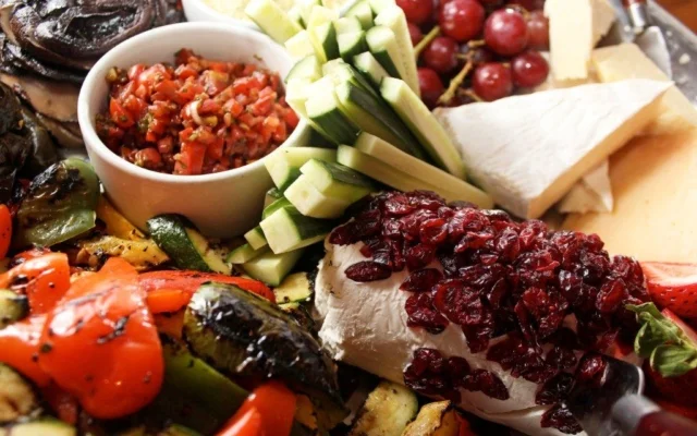 A cheese platter with grapes, celery sticks, cherry tomatoes, a small bowl of salsa, and various cheeses, some topped with cranberries.